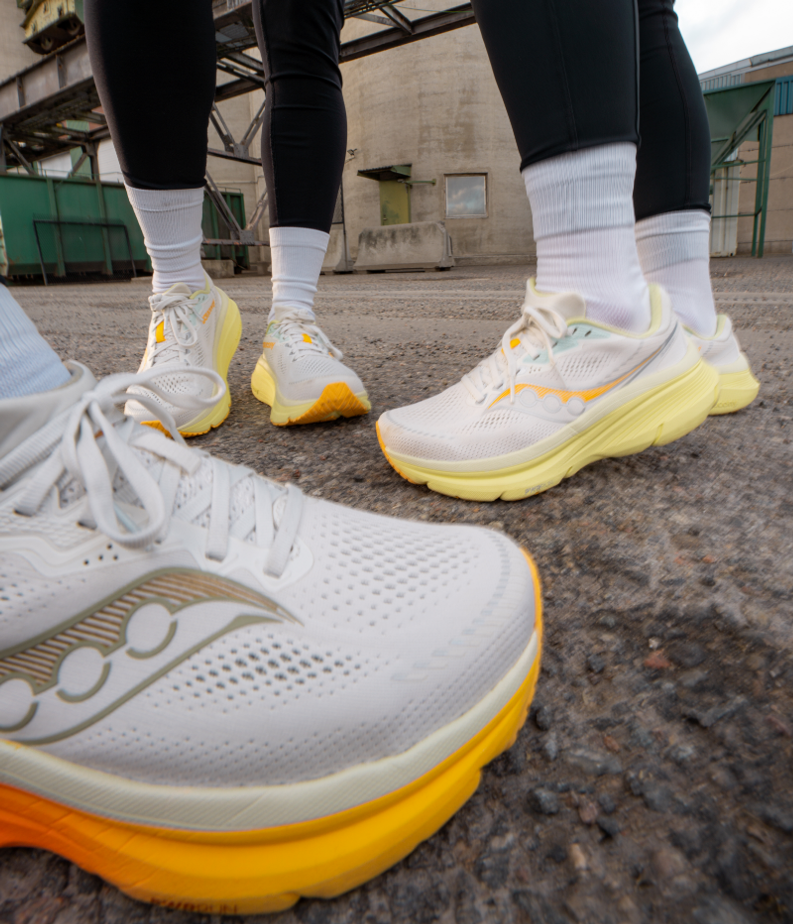 Three pairs of white Saucony running shoes with yellow and orange soles on asphalt, with an industrial building in the background.