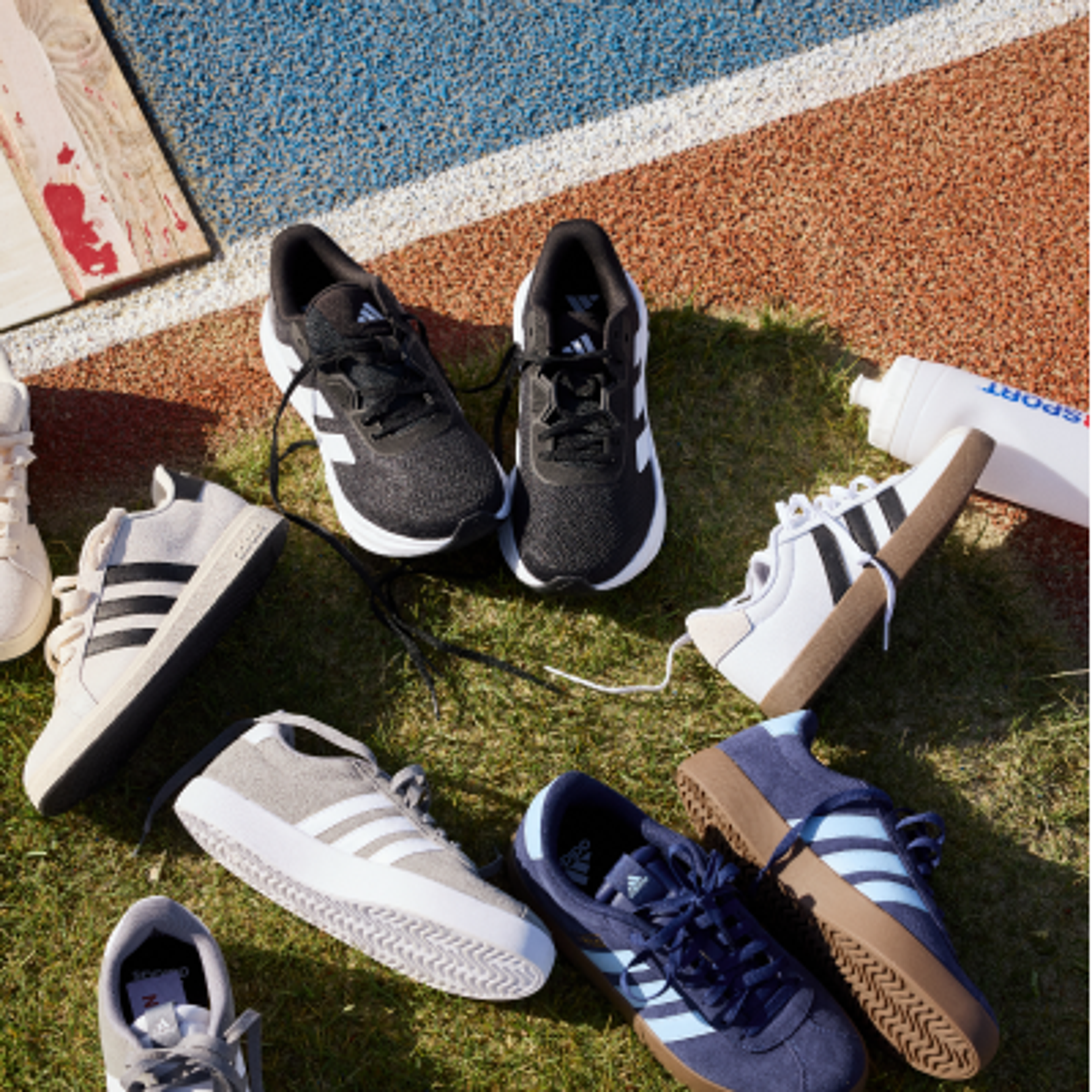 Multiple pairs of Adidas athletic shoes on green grass next to a track, with a white water bottle.