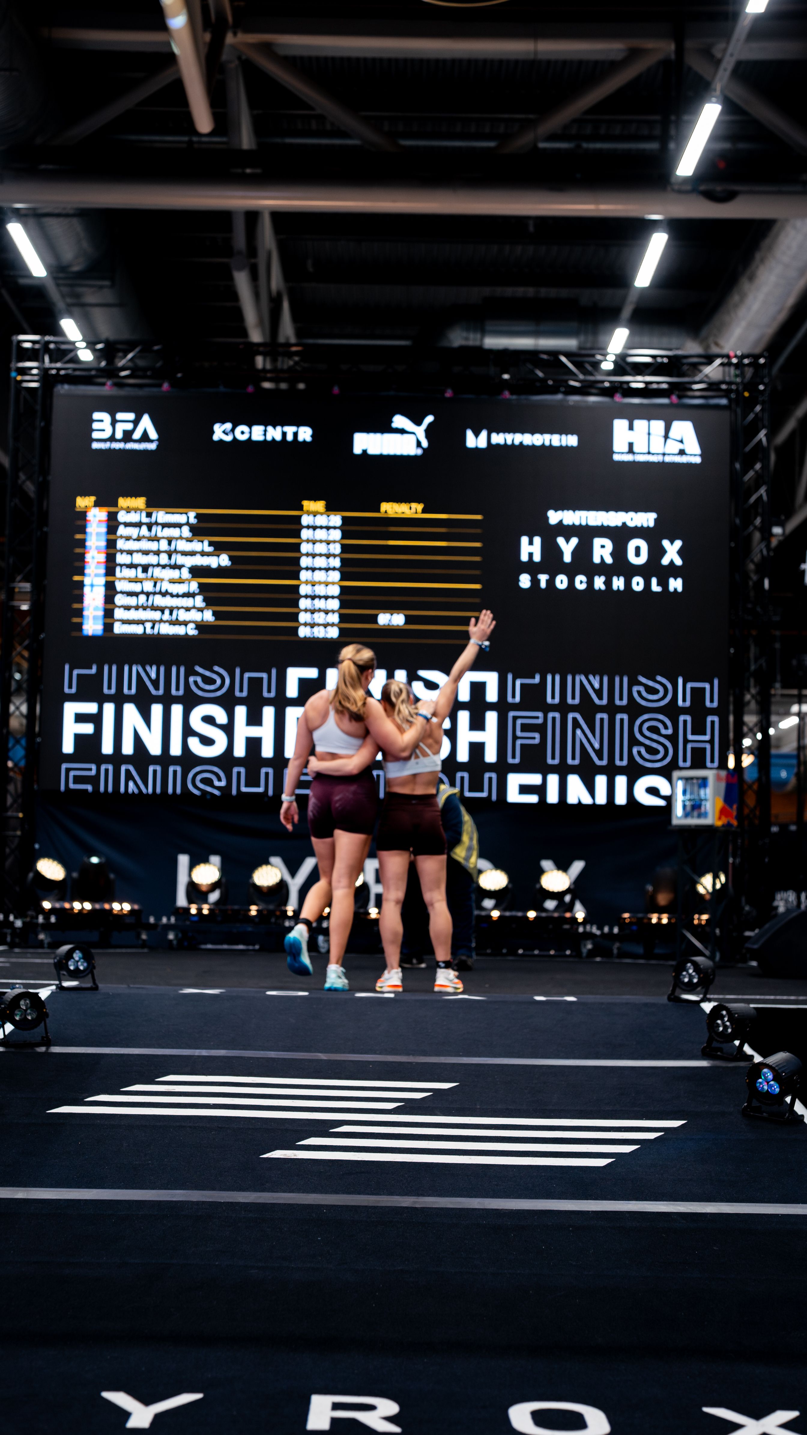 Two athletes hug on a race floor, with a large screen showing Hyrox results and "FINISH" behind them.