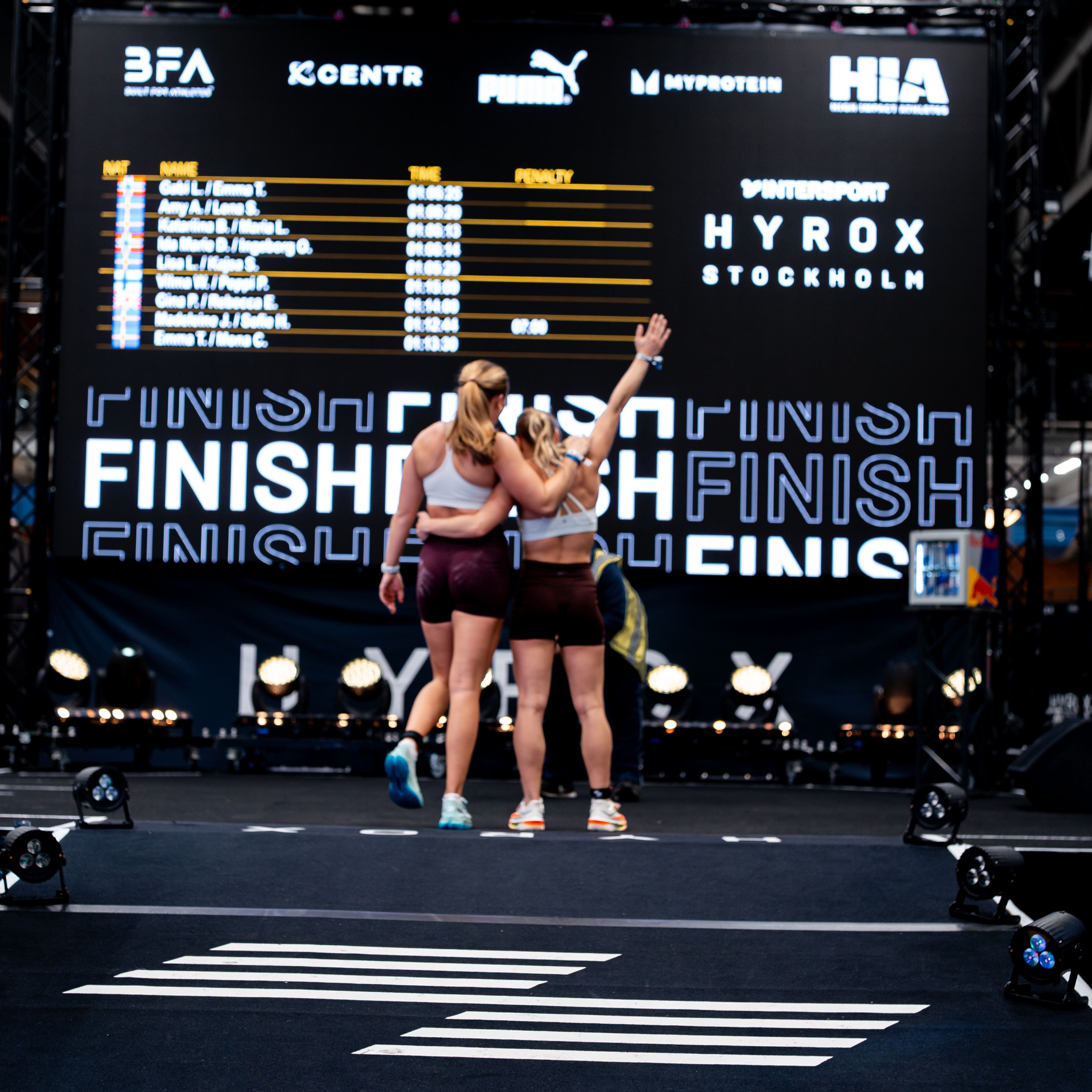 Two athletes hug on a race floor, with a large screen showing Hyrox results and "FINISH" behind them.