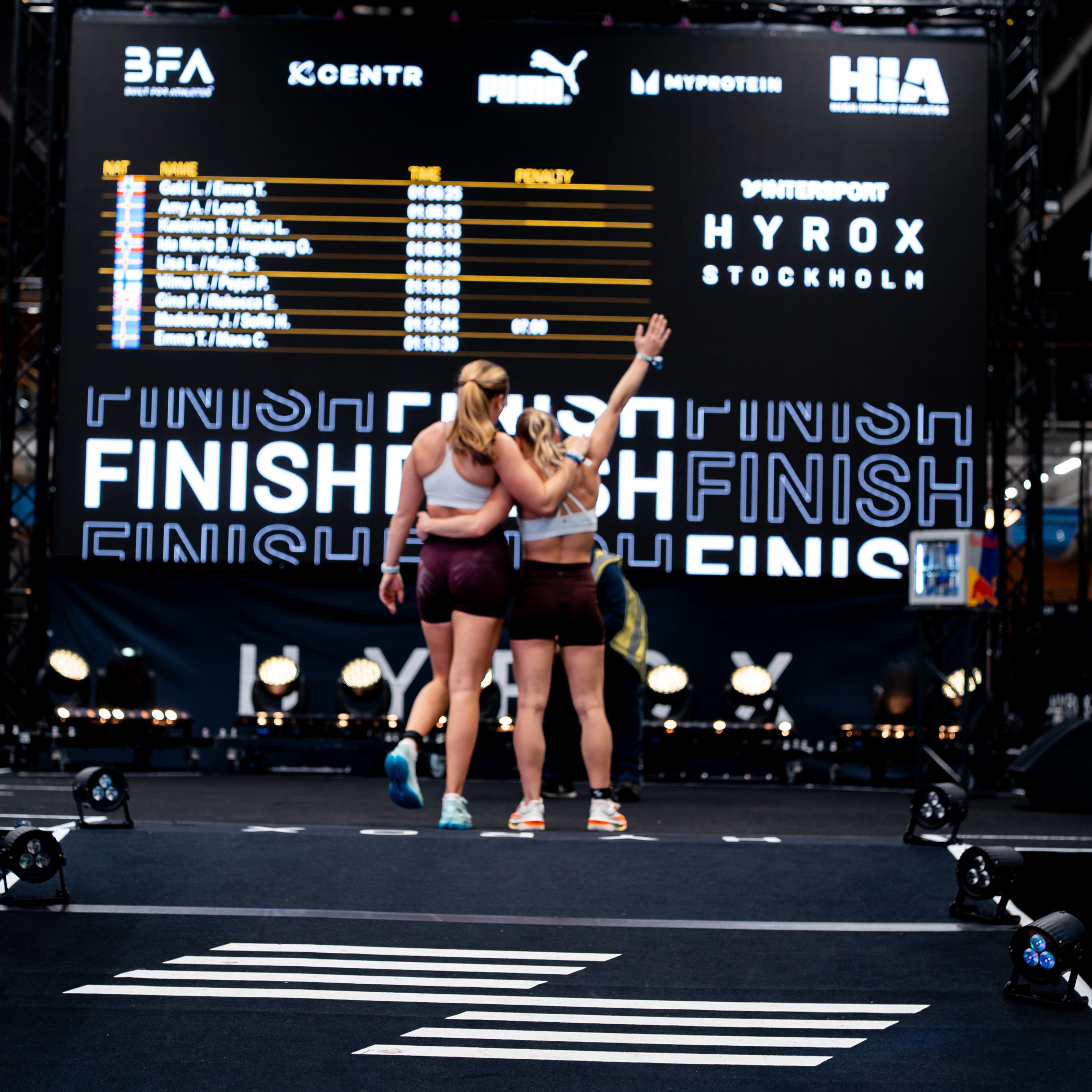 Two athletes hug on a race floor, with a large screen showing Hyrox results and "FINISH" behind them.