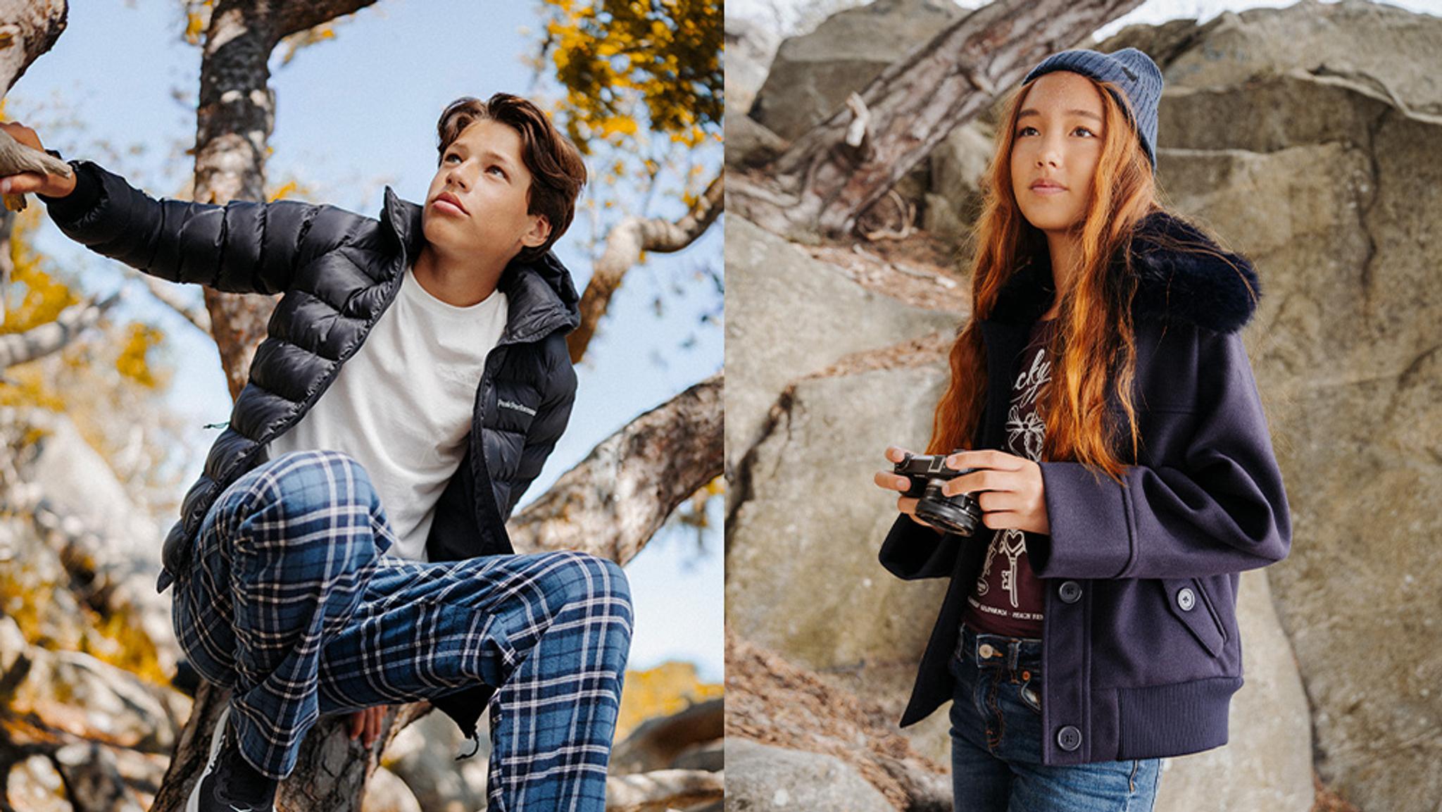 Boy in a black puffer jacket climbing a tree, and a girl in a navy coat holding a camera, both outdoors.
