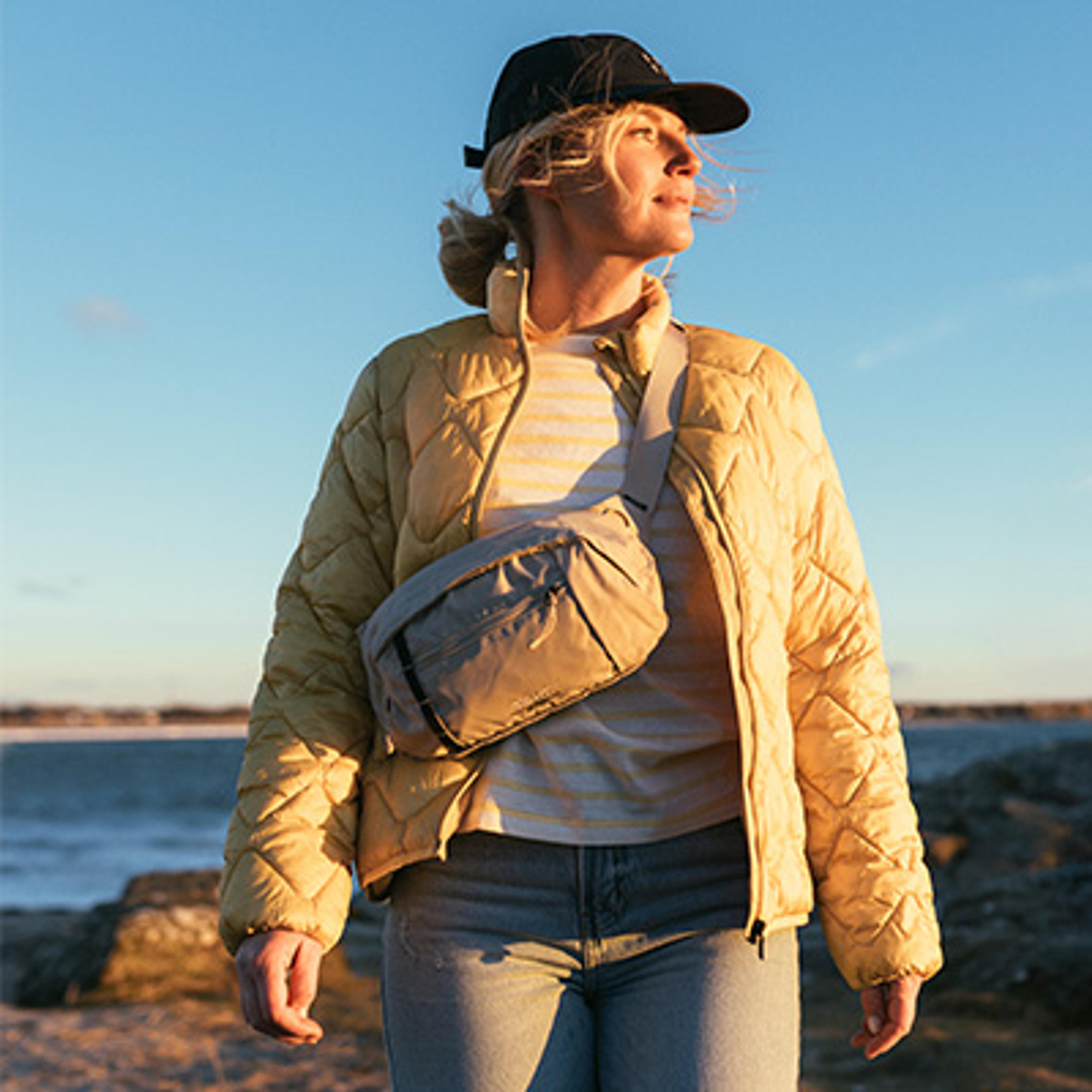 A woman in a black cap, yellow quilted jacket, and grey cross-body bag looks out at a coastline under a blue sky.
