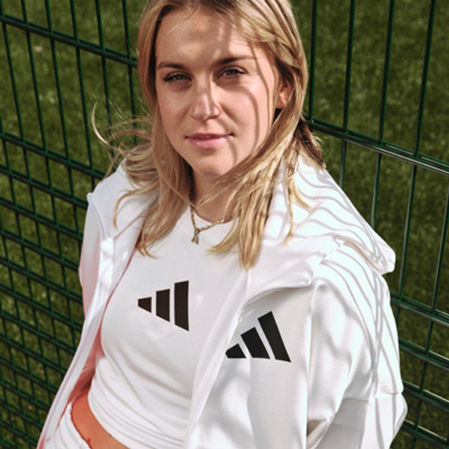a woman wearing a white adidas jacket is standing next to a fence .