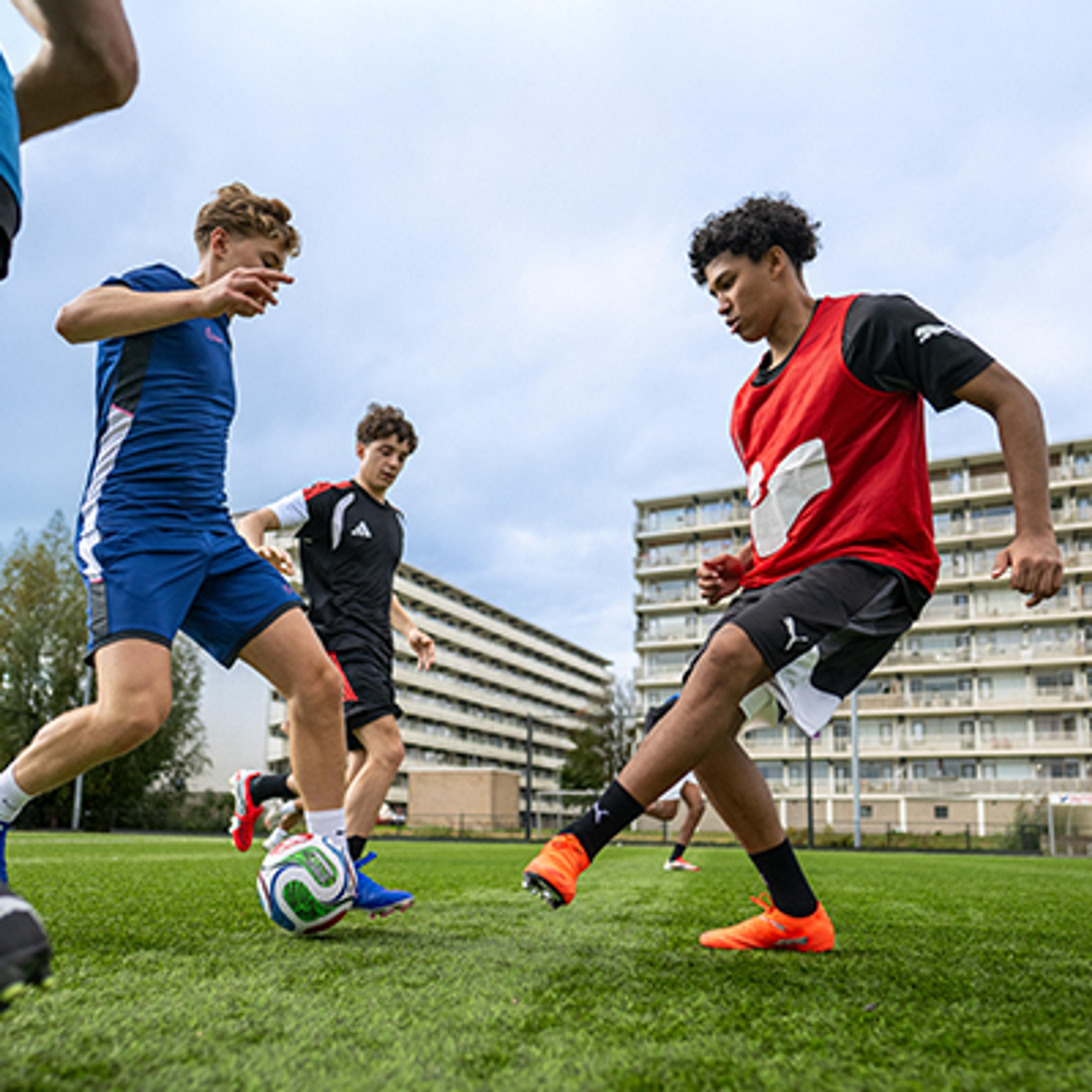 Young men playing soccer on a green field with apartment buildings in the background.