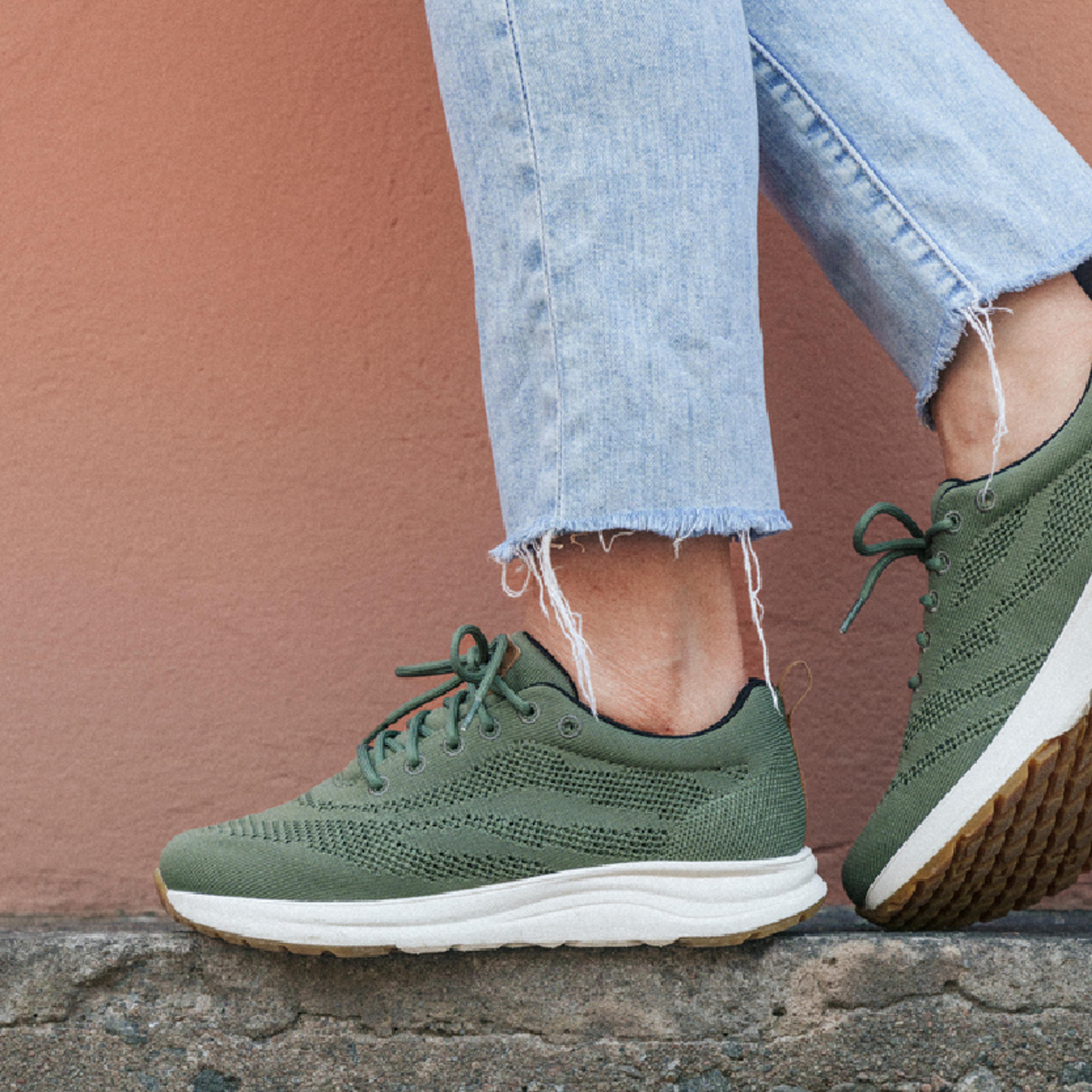 A person wears light blue jeans with frayed hems and green knit sneakers while standing on a concrete ledge.