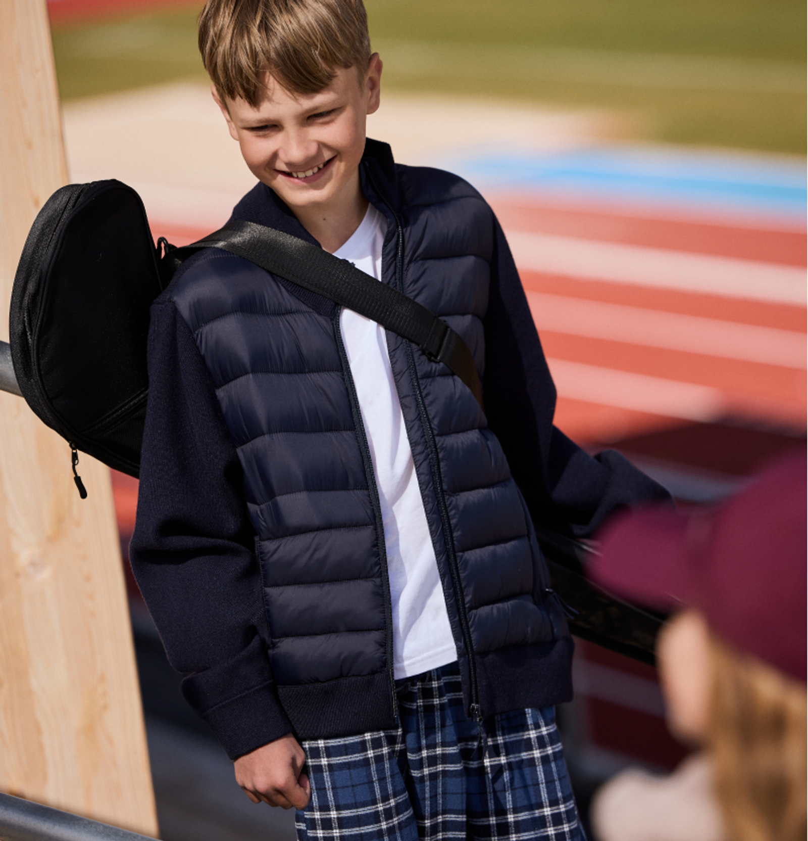 A smiling boy in a navy puffer jacket and plaid pants with a black bag over his shoulder.