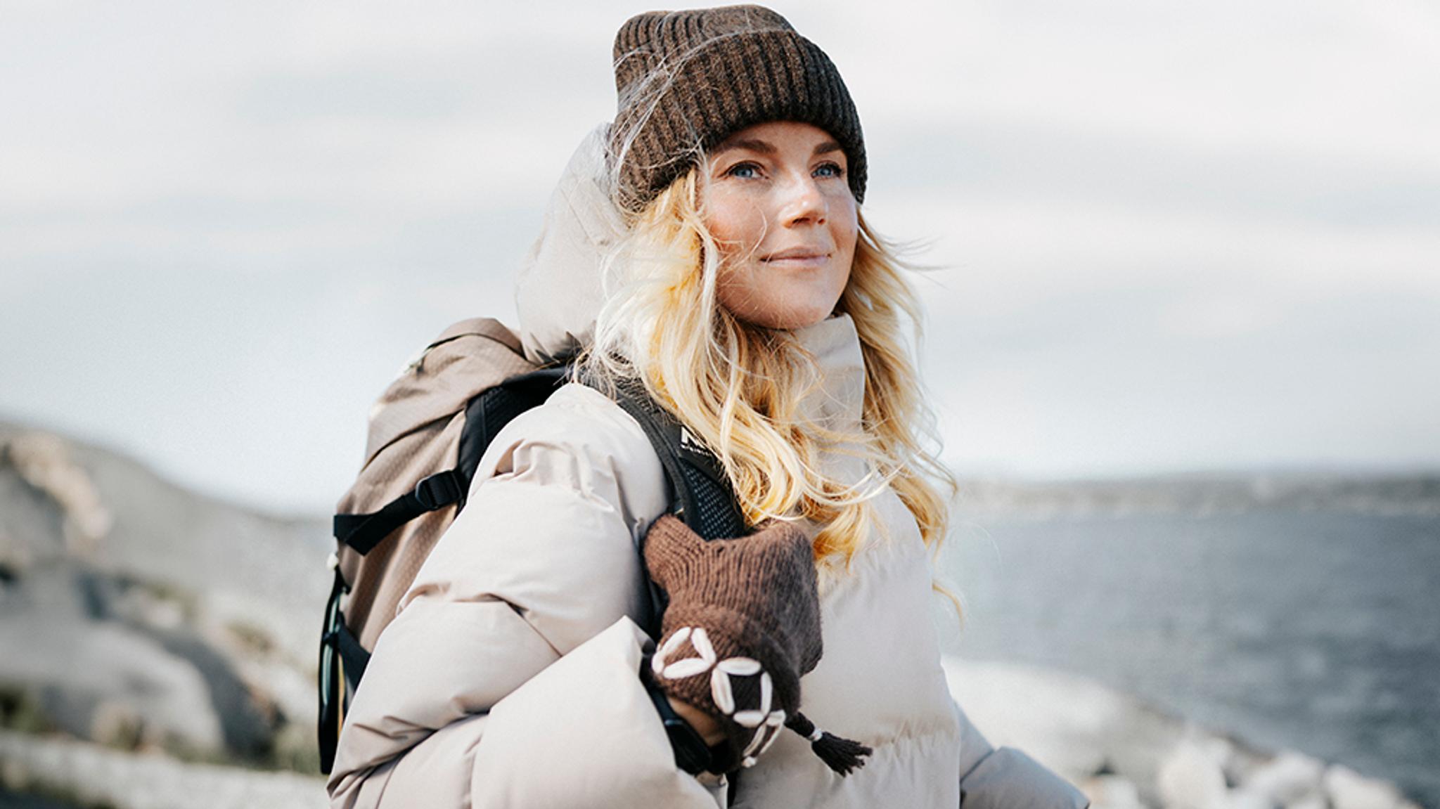 A blonde woman in a beanie, light puffer jacket, and backpack, wearing a patterned brown mitten, looks calmly towards the right with water in the background.