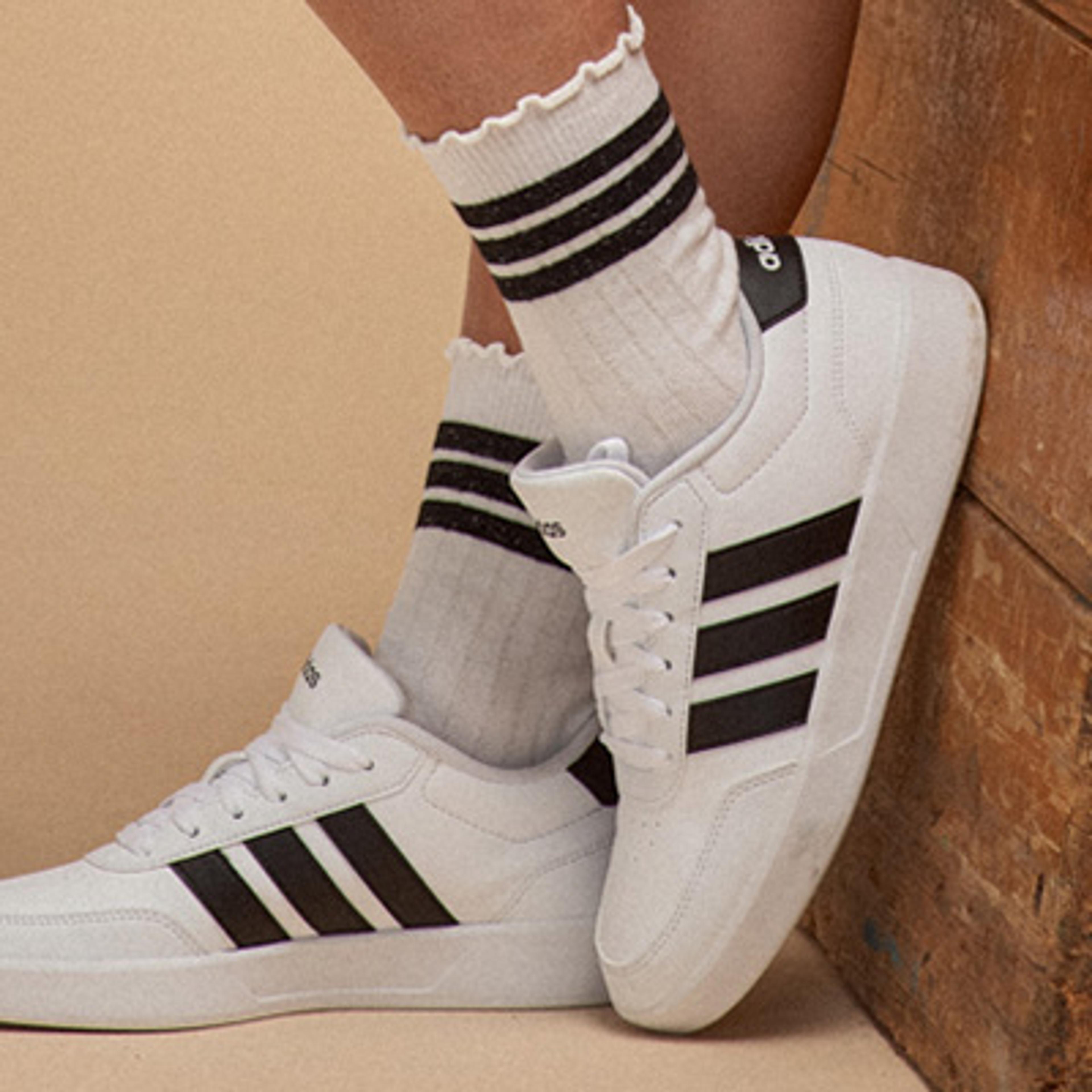 Close-up of feet wearing white Adidas sneakers with black stripes and white ribbed socks, leaning against wood.