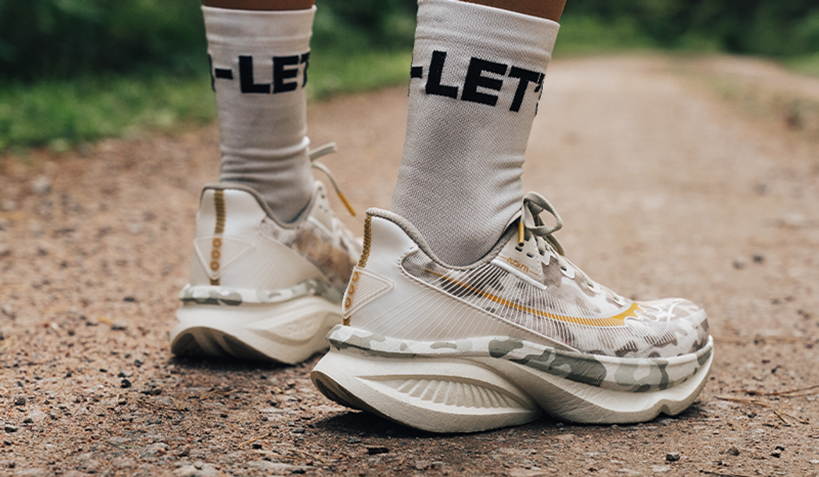 Feet wearing white socks and camouflage running shoes with thick soles on a dirt path.