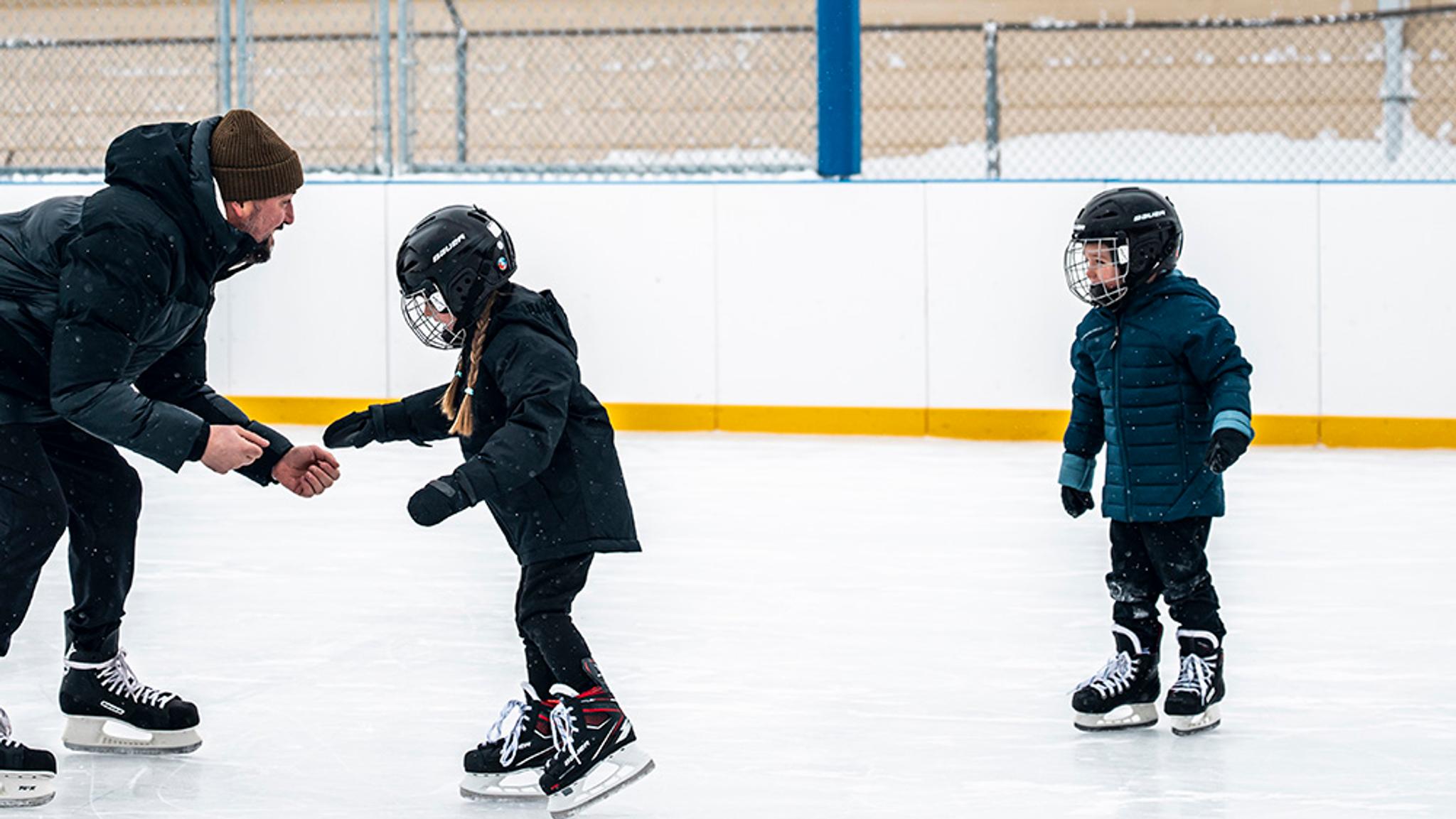 An adult helps a young girl in a helmet learn to ice skate, with another child standing nearby on the rink.