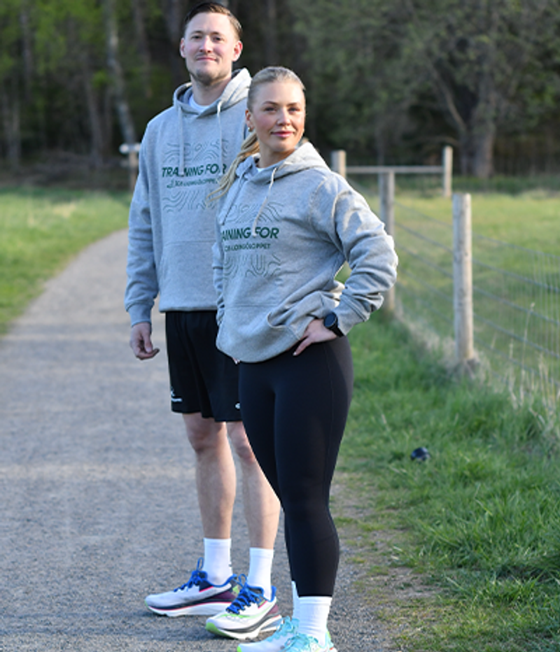 A man and a woman in grey "TRAINING FOR LÖPLÖRDAGLOPPET" hoodies stand on a path outdoors.