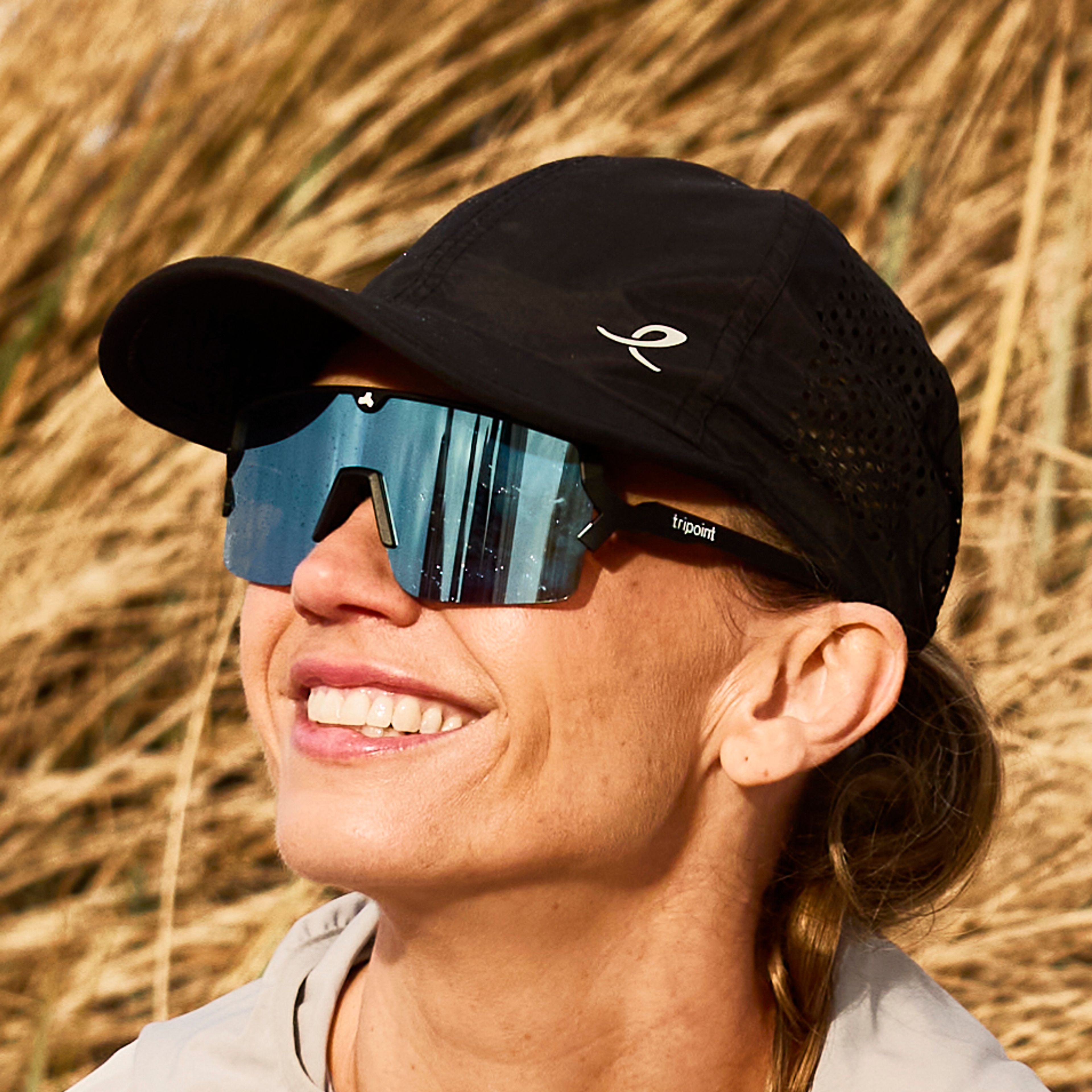 A woman in a cap and sunglasses stretches on a sandy beach in front of dry dune grass.