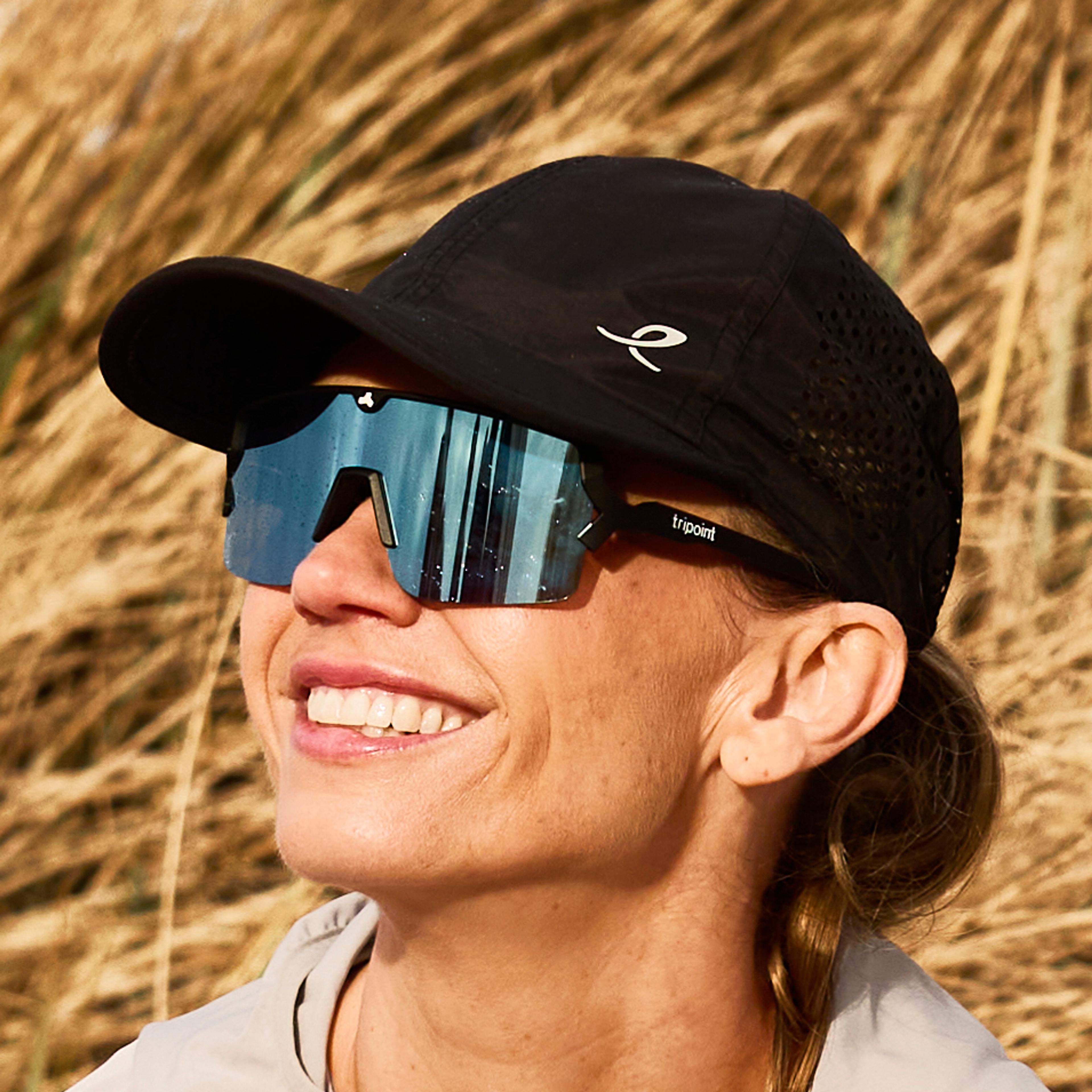 A woman in a cap and sunglasses stretches on a sandy beach in front of dry dune grass.