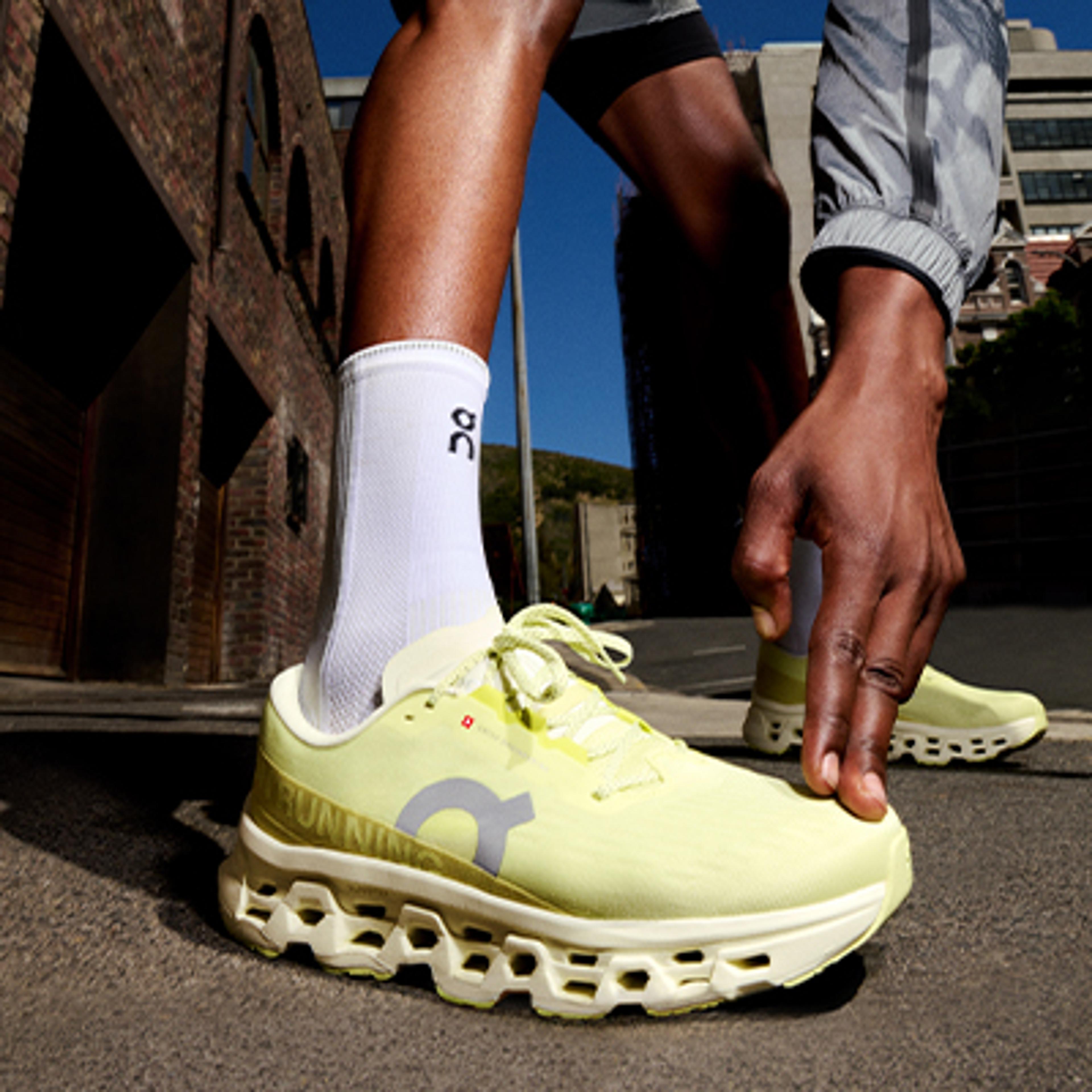 A person wearing bright yellow On Running shoes and white socks bends to adjust a shoe on an urban street.