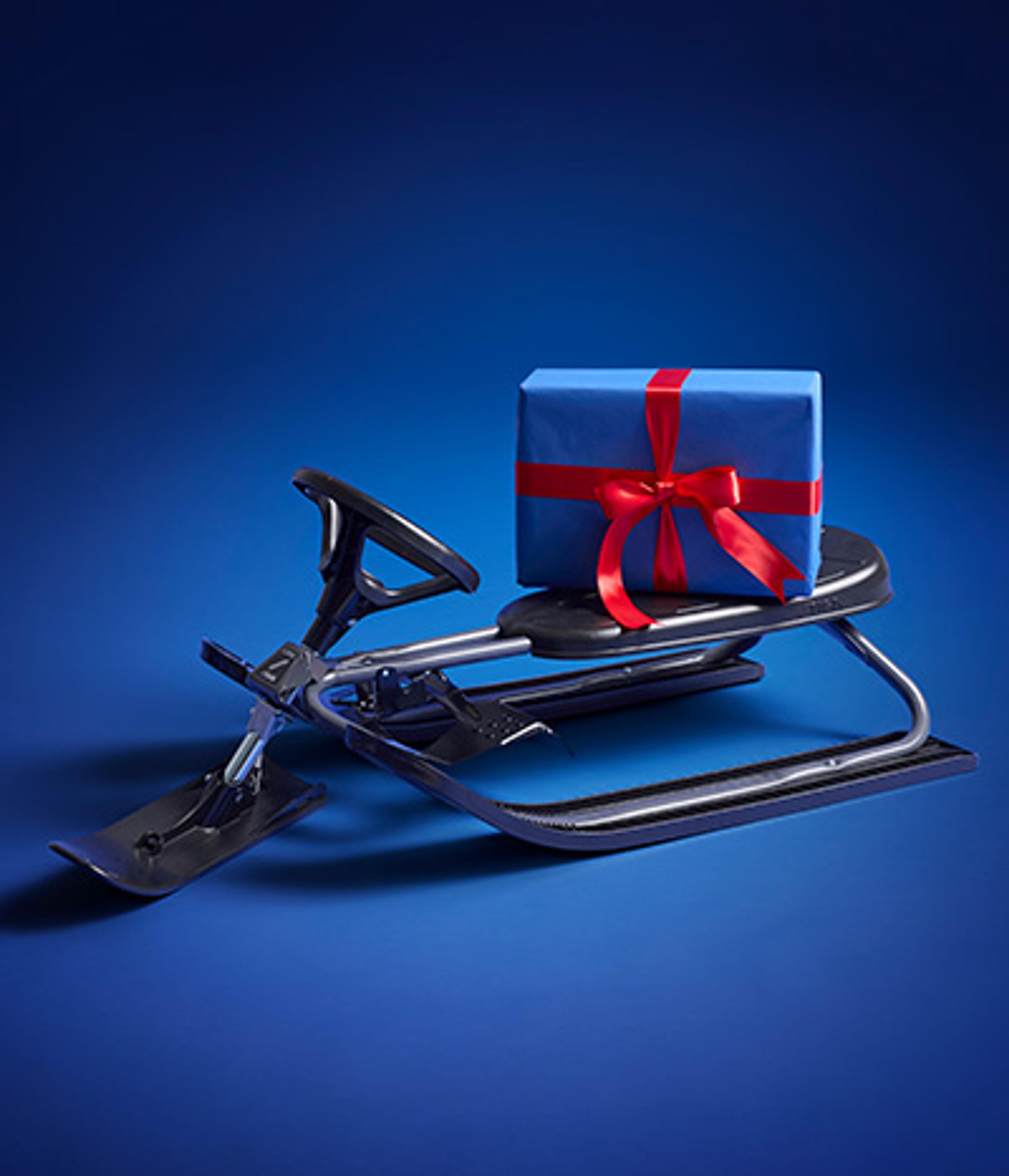 A blue gift box with a red ribbon rests on a dark gray snow racer sled, on a blue background.