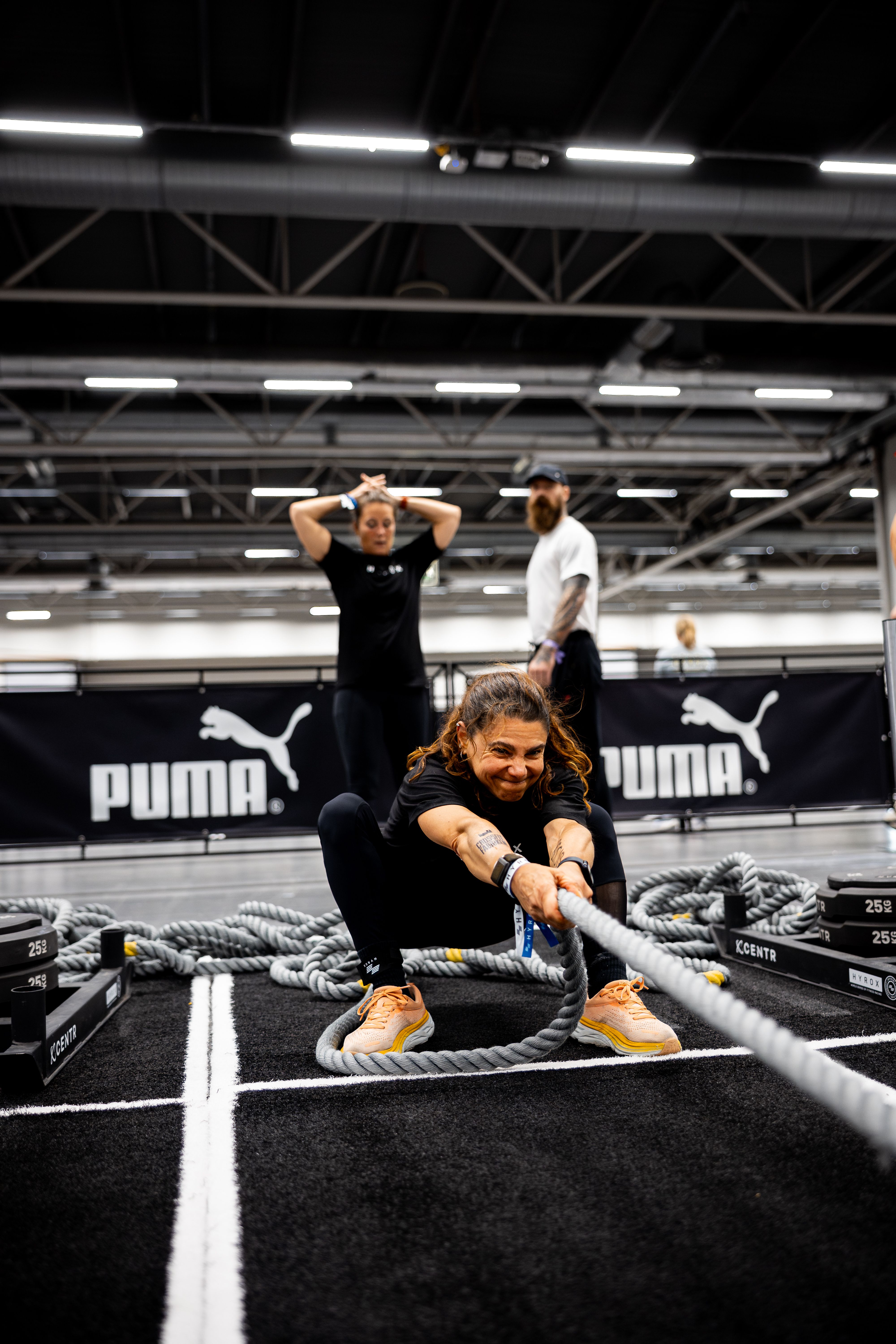 A woman crouches, pulling a thick rope with a determined expression, while two people stand behind her near a PUMA banner.