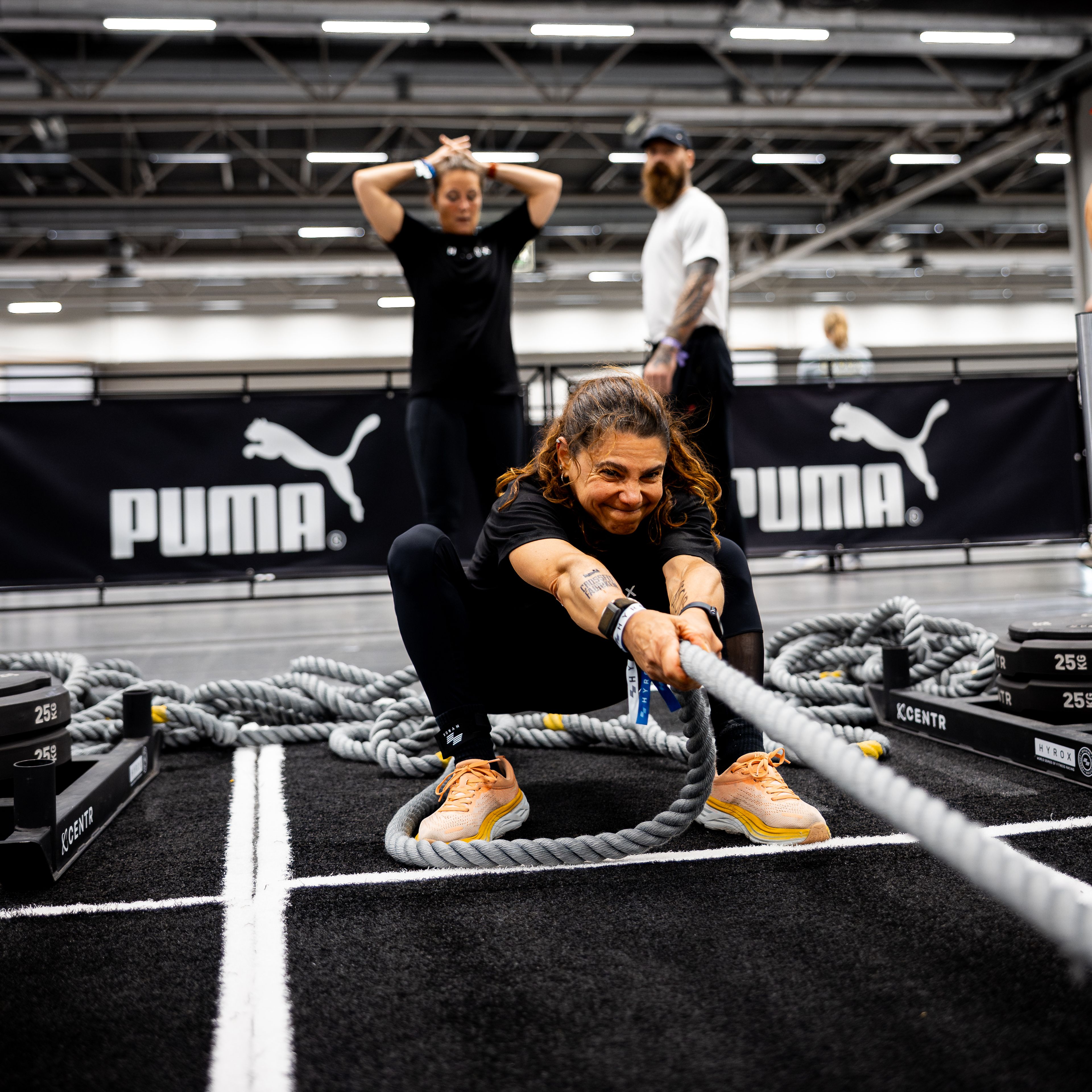 A woman crouches, pulling a thick rope with a determined expression, while two people stand behind her near a PUMA banner.