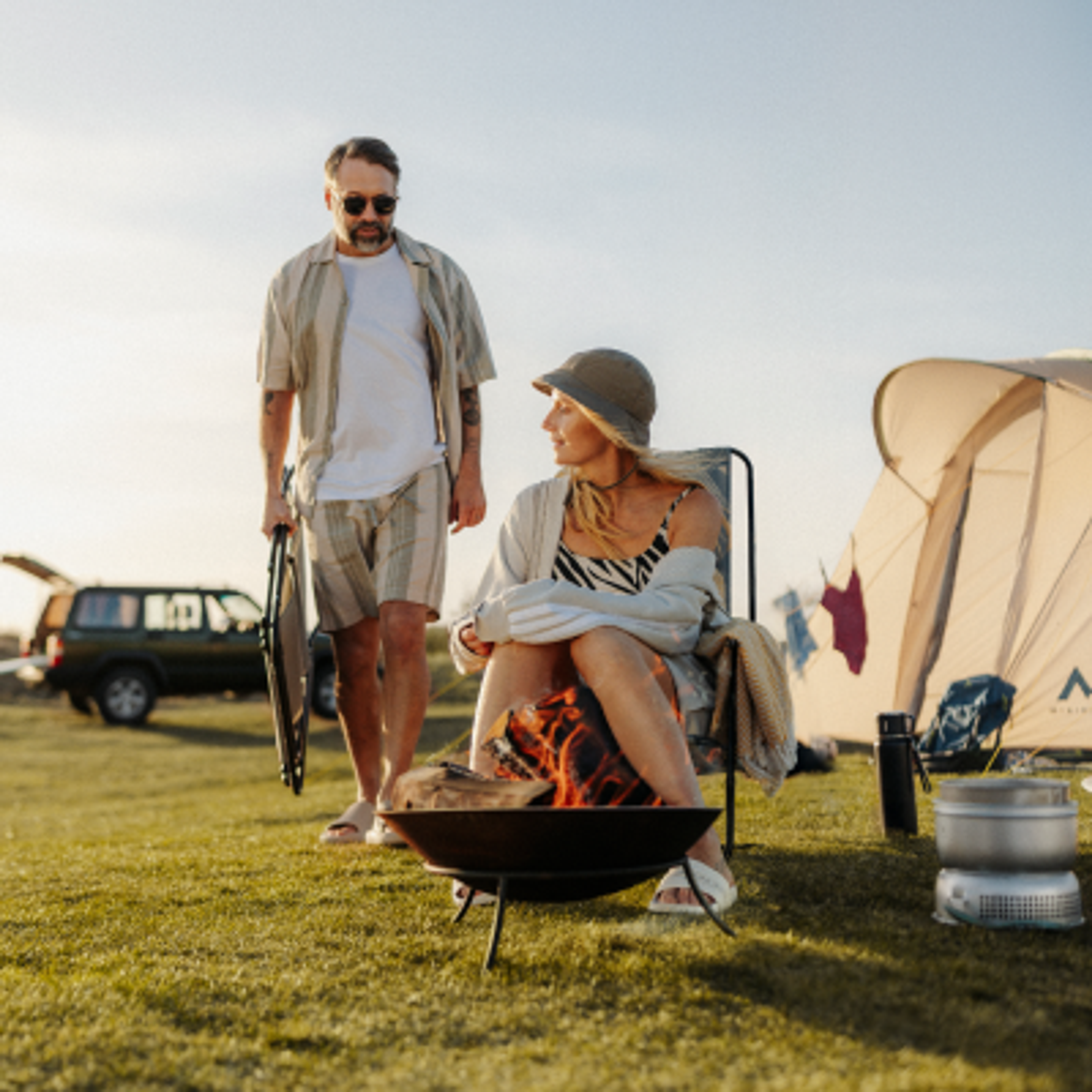 A couple camping with a campfire, tent, and car in a grassy field.