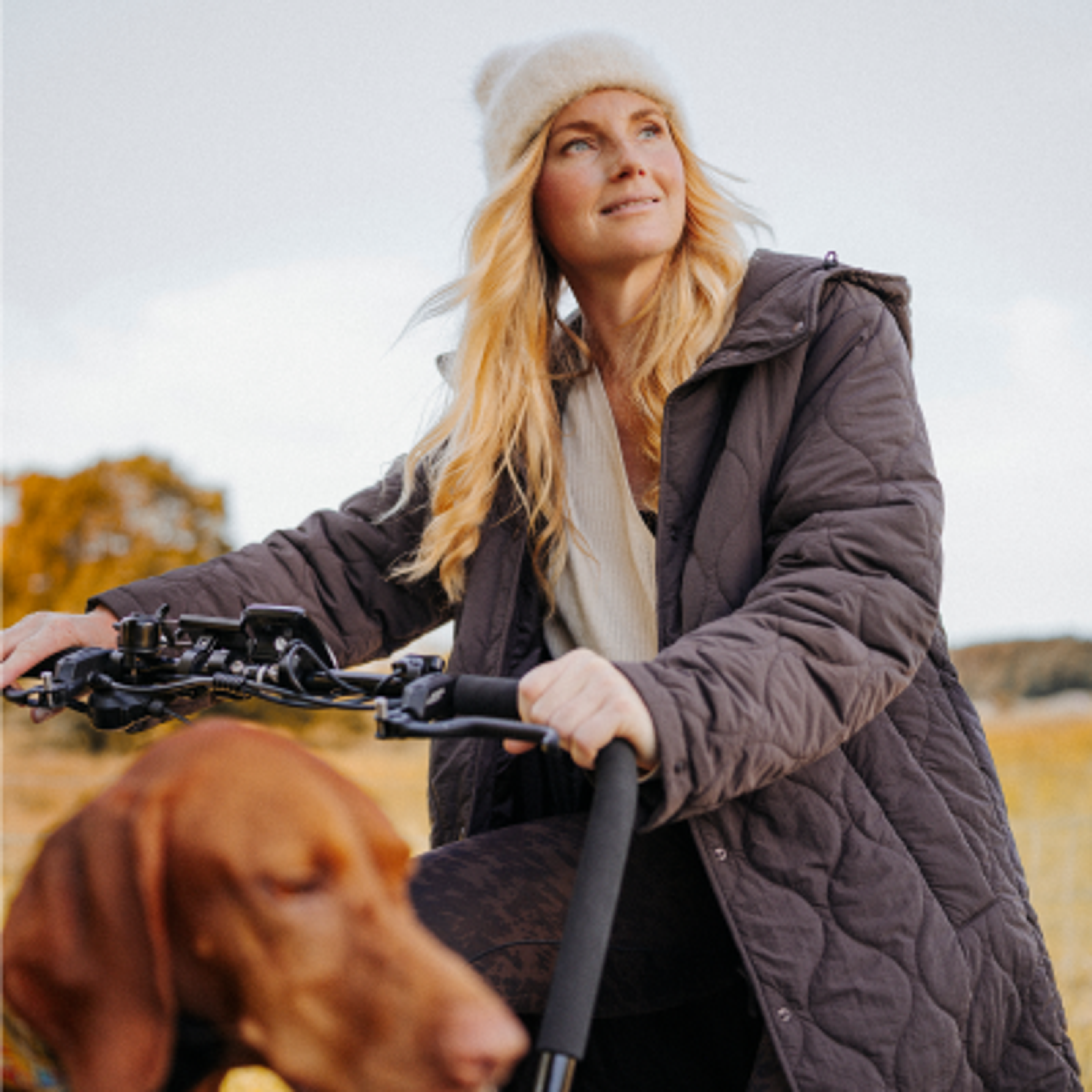 A woman in a beanie and quilted jacket on a bike with her dog, looking up at an autumn sky.