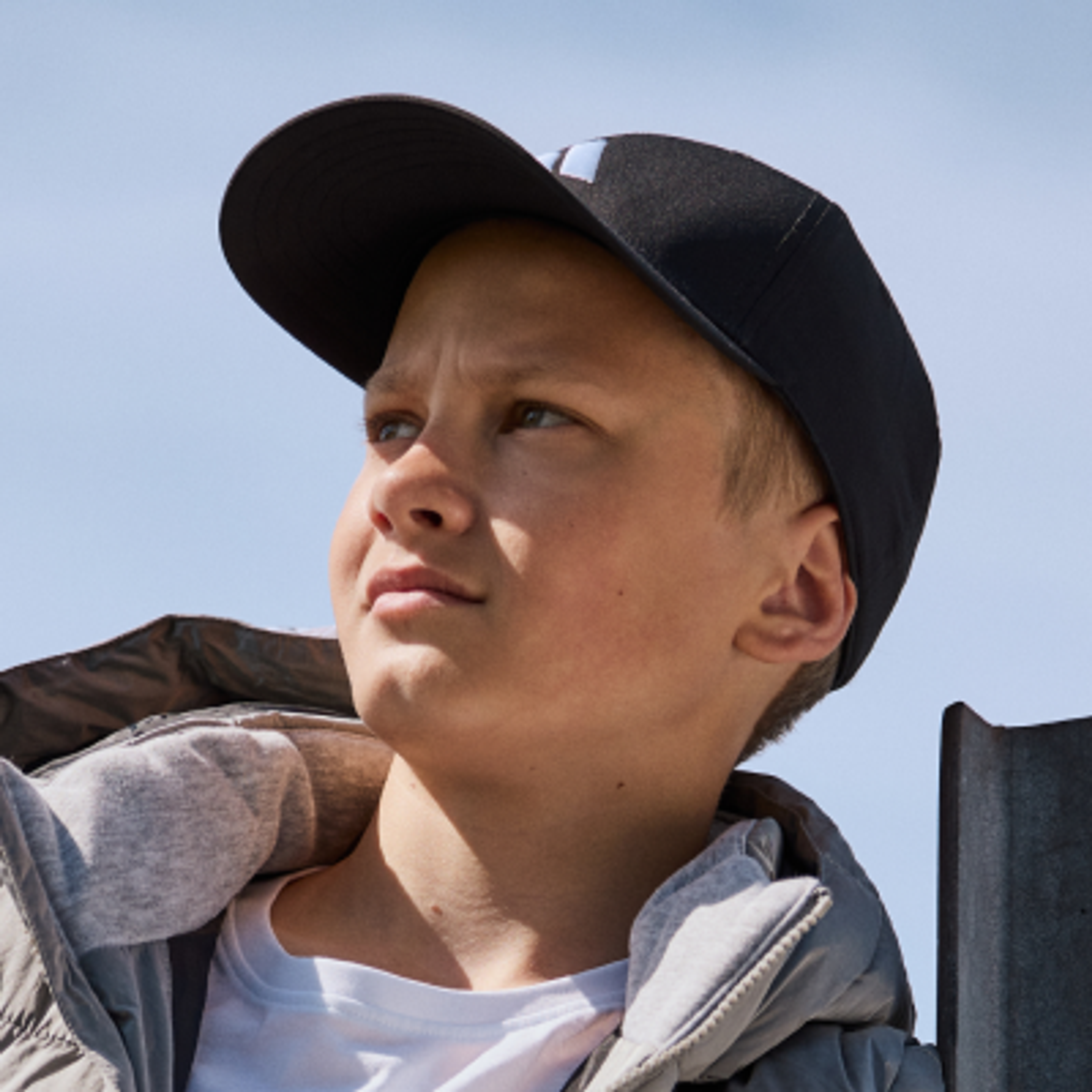 A young boy in a black cap and grey jacket looks up against a blue sky.