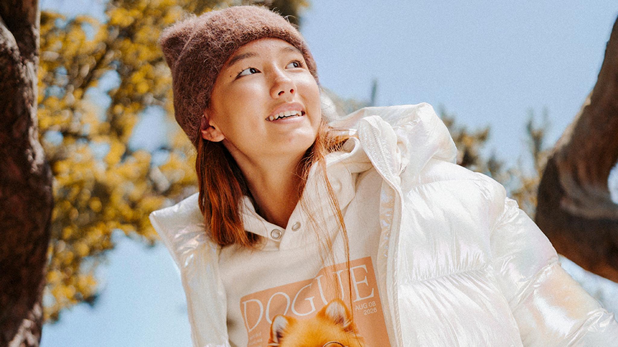A young woman in a brown beanie and iridescent white puffer jacket smiles while looking up outdoors.