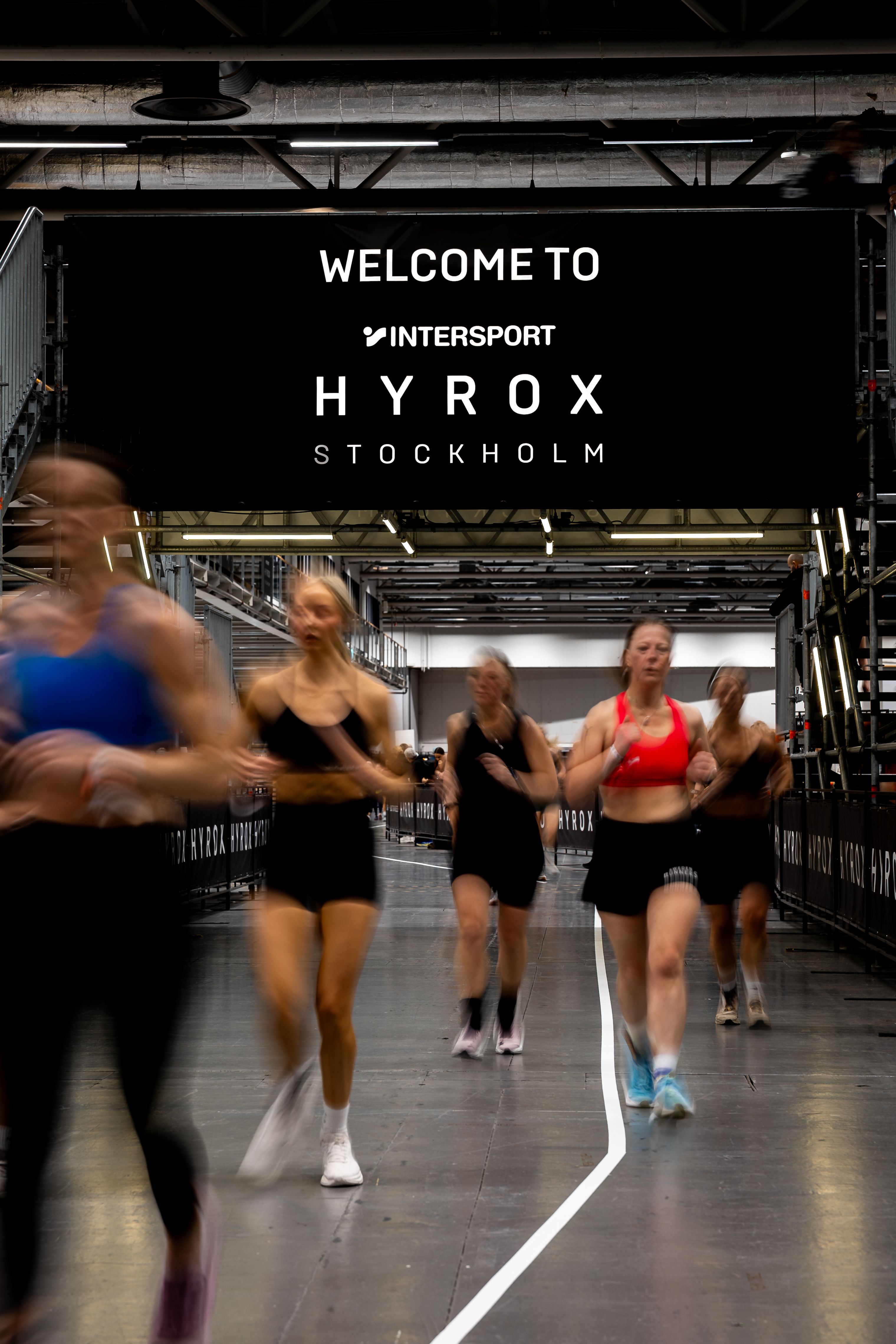 Blurred women running at an indoor HYROX Stockholm event, under a banner reading 'WELCOME TO INTERSPORT HYROX STOCKHOLM'.