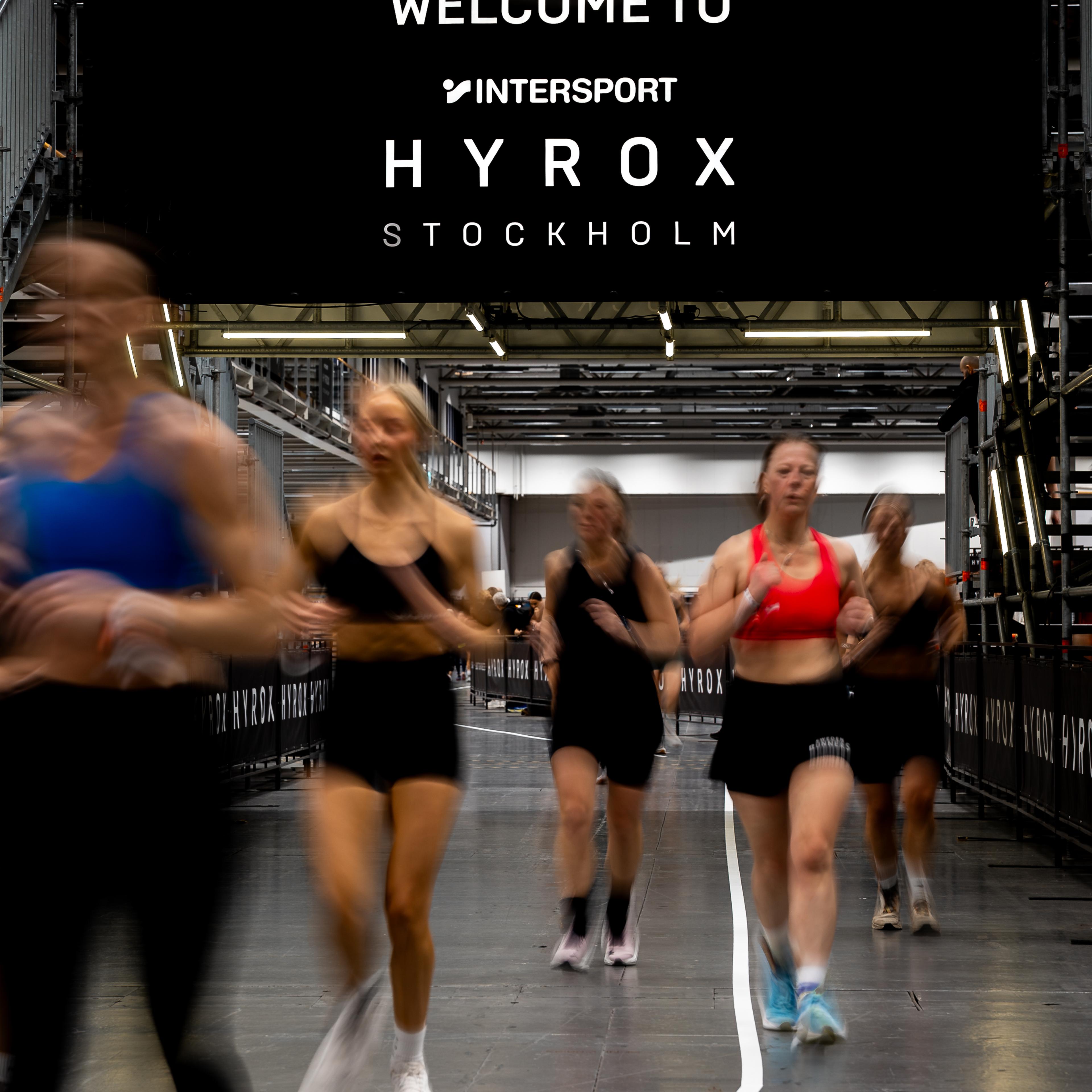 Blurred women running at an indoor HYROX Stockholm event, under a banner reading 'WELCOME TO INTERSPORT HYROX STOCKHOLM'.