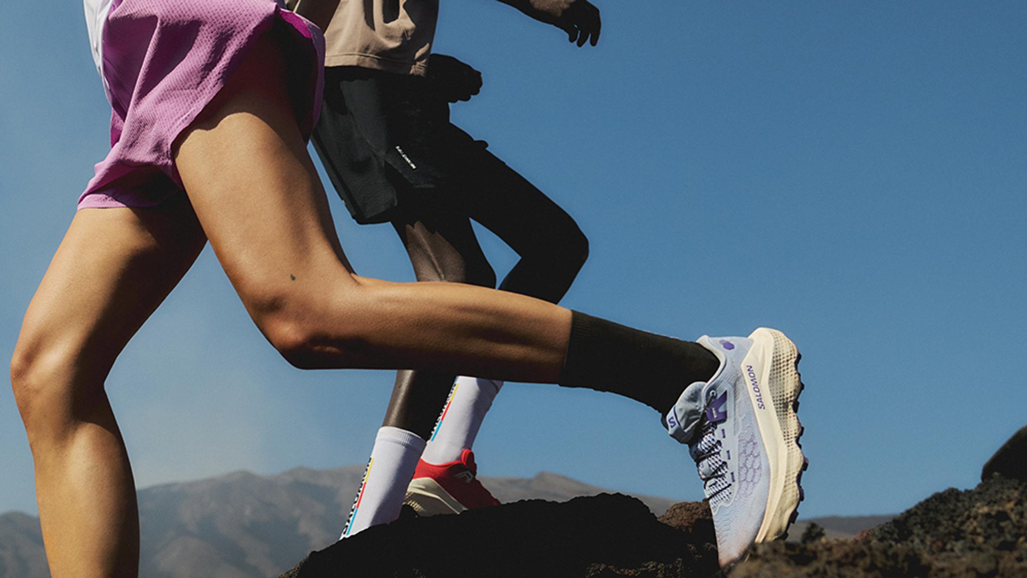 Two runners' legs in motion on a rocky trail, one wearing lavender trail shoes, under a blue sky.