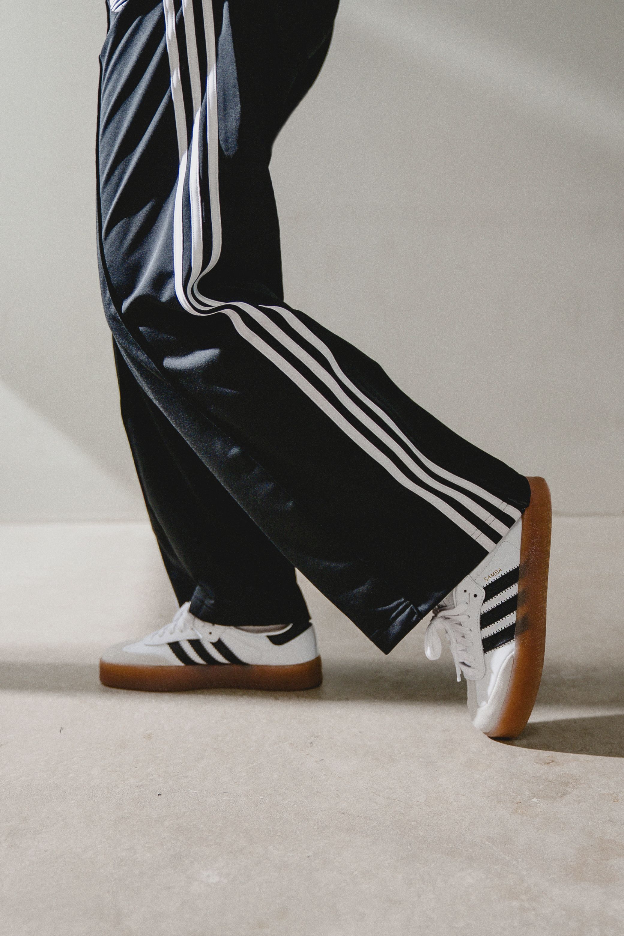 Person wearing black Adidas track pants with white stripes and white Adidas Samba sneakers with gum soles.