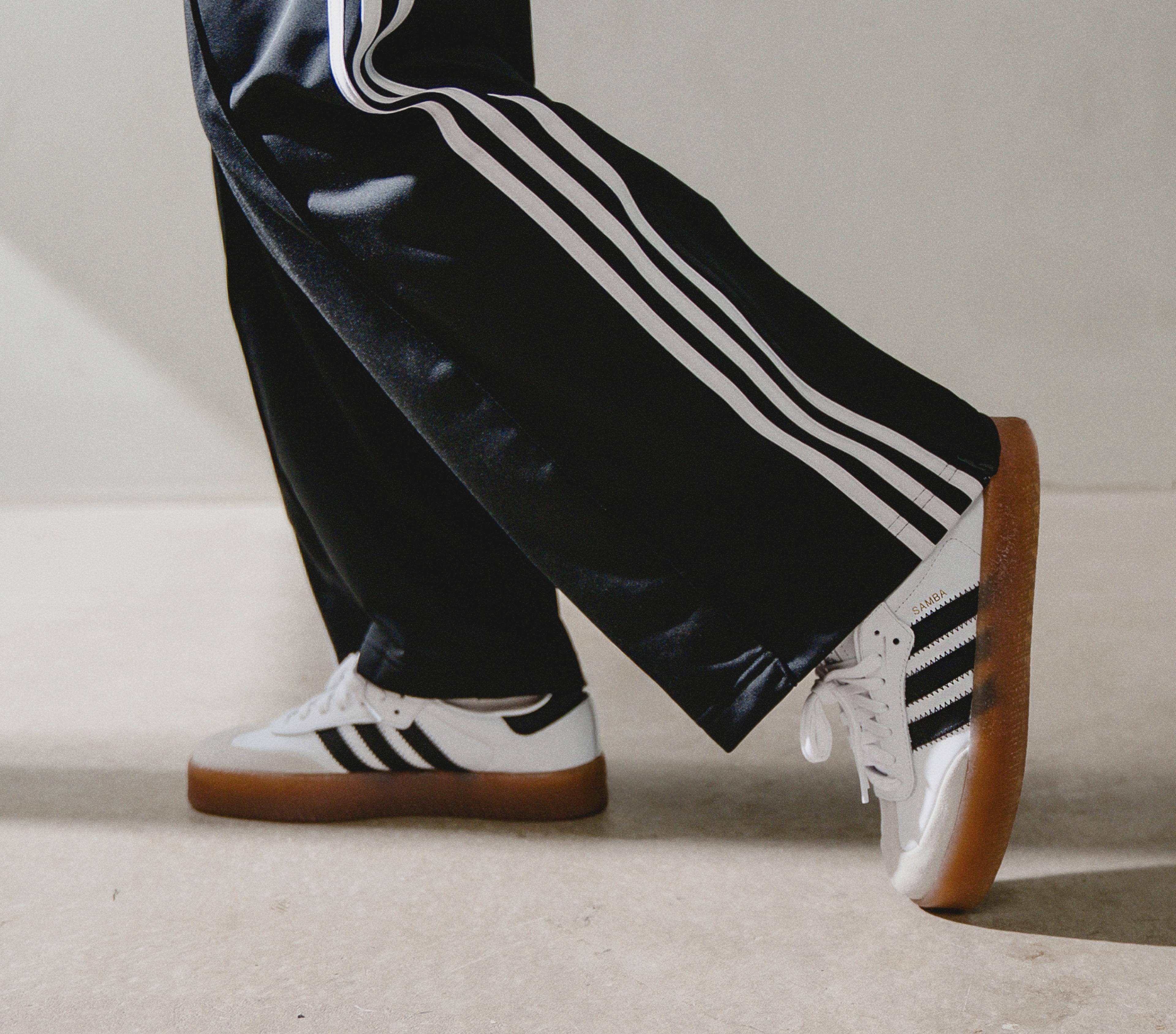 Person wearing black Adidas track pants with white stripes and white Adidas Samba sneakers with gum soles.