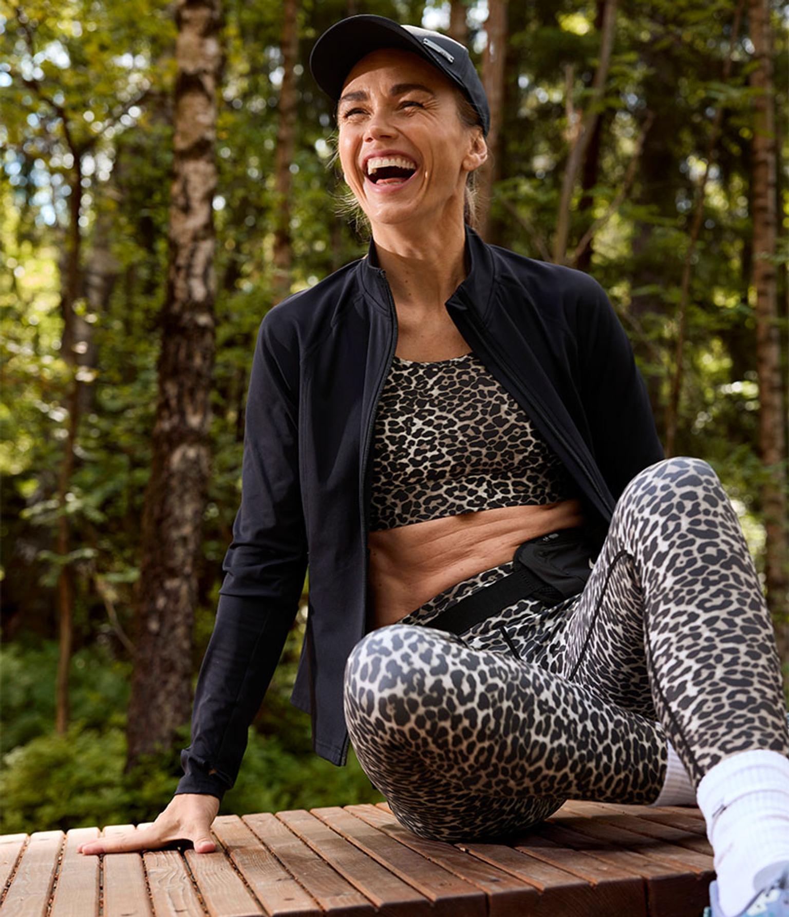 a woman is sitting on a wooden bench in the woods wearing leopard print leggings and a black jacket .
