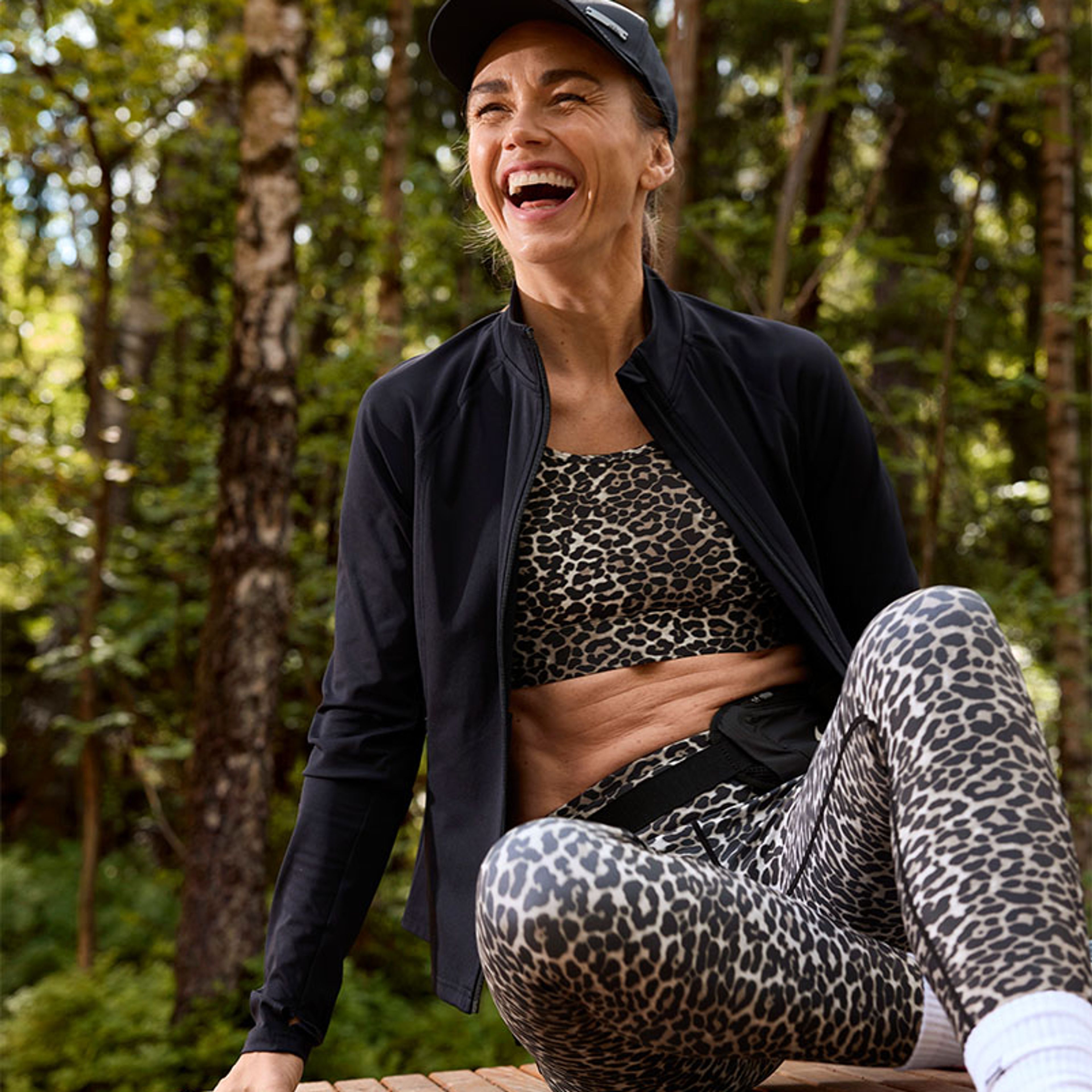 a woman is sitting on a wooden bench in the woods wearing leopard print leggings and a black jacket .