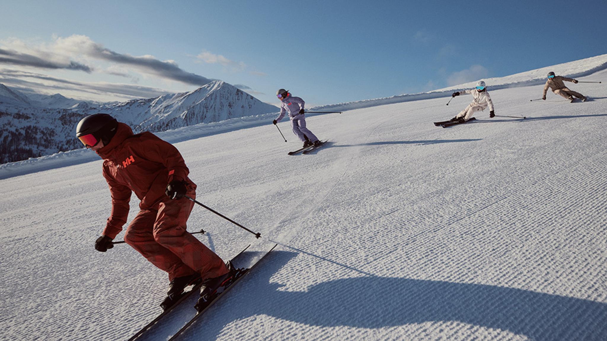 Four people skiing down a sunlit, groomed ski slope with mountains in the background.
