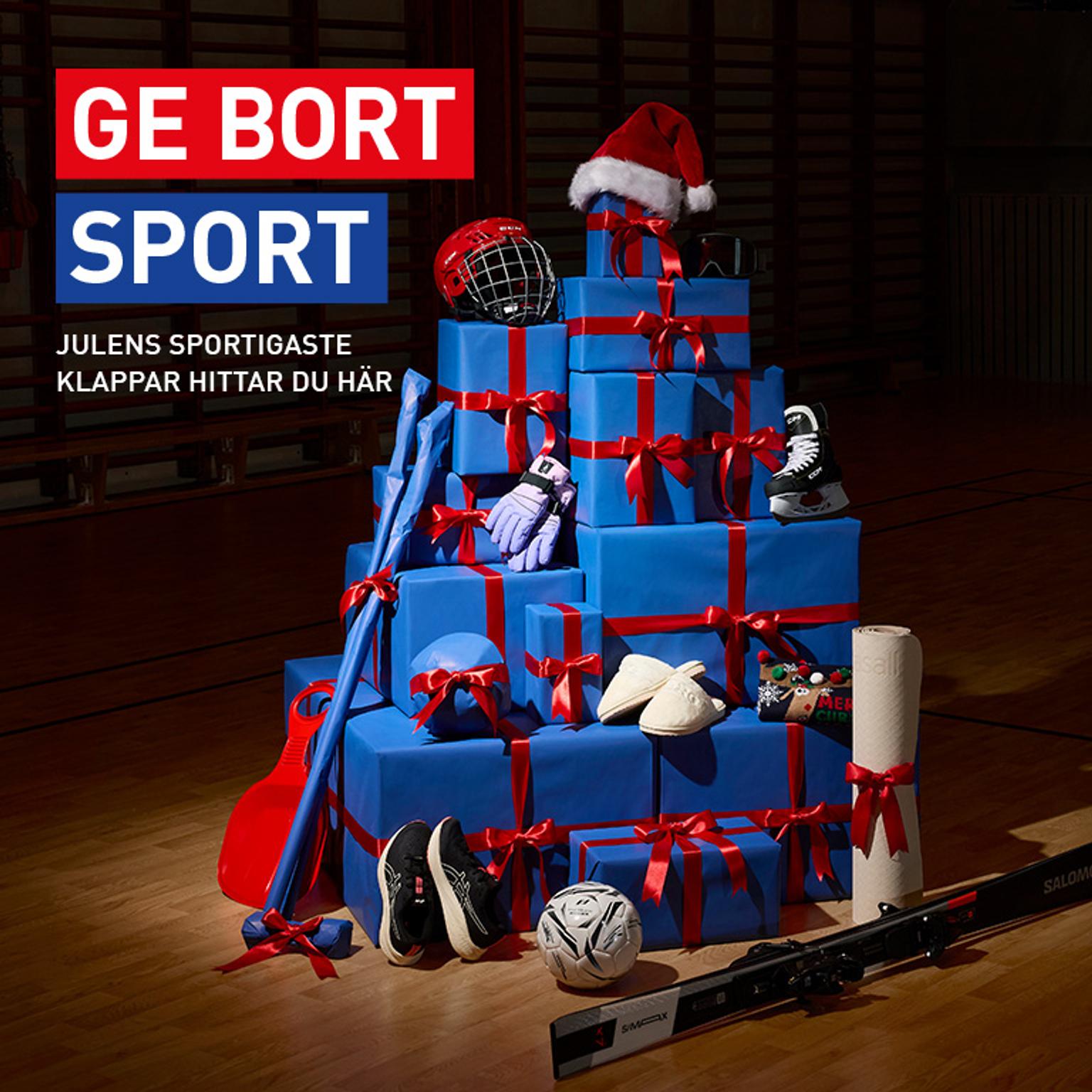 A large stack of blue gift boxes with red ribbons, surrounded by various sports equipment, in a gym. Text: "GE BORT SPORT."