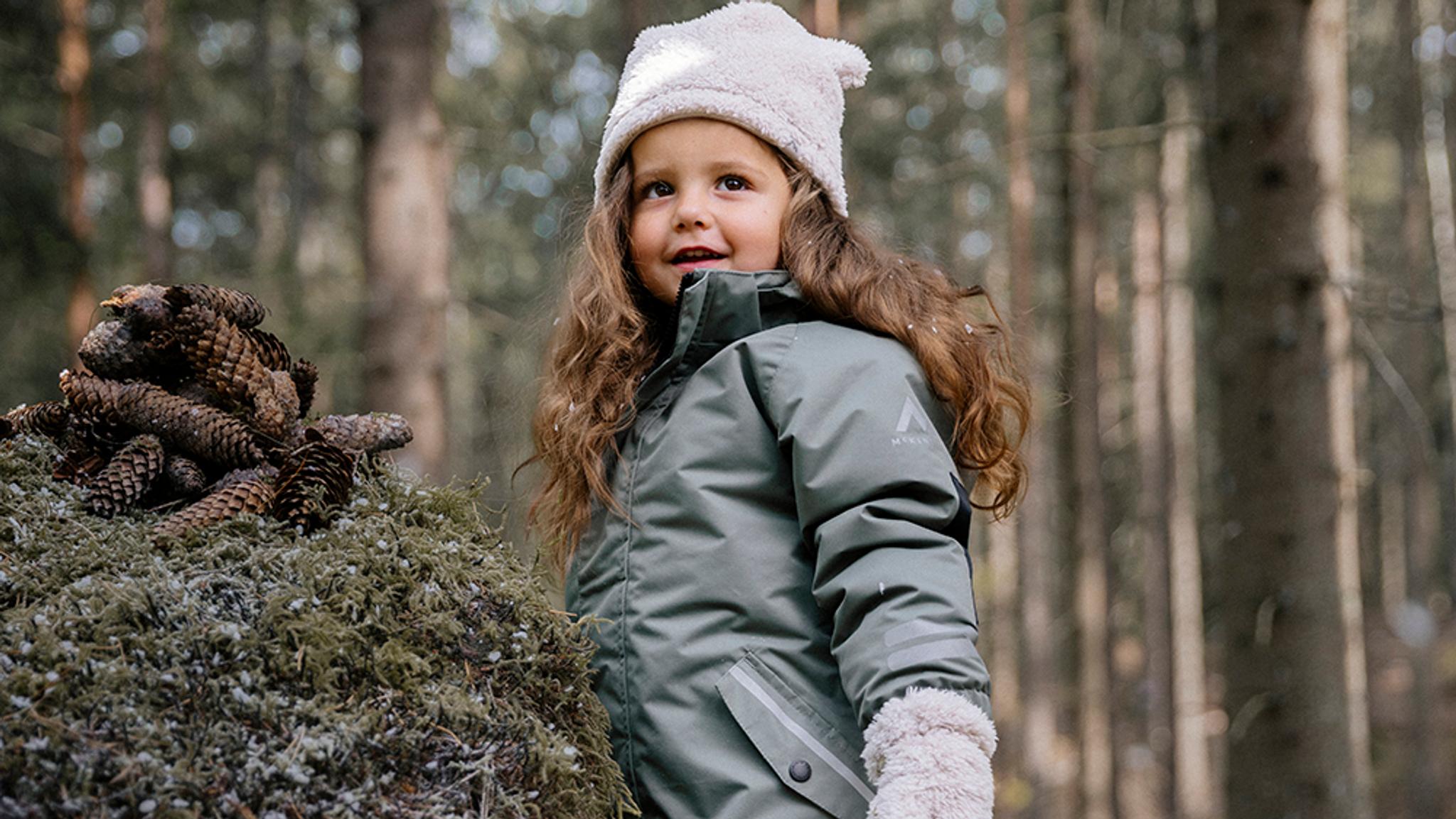 a little girl is standing next to a pile of pine cones in the woods .