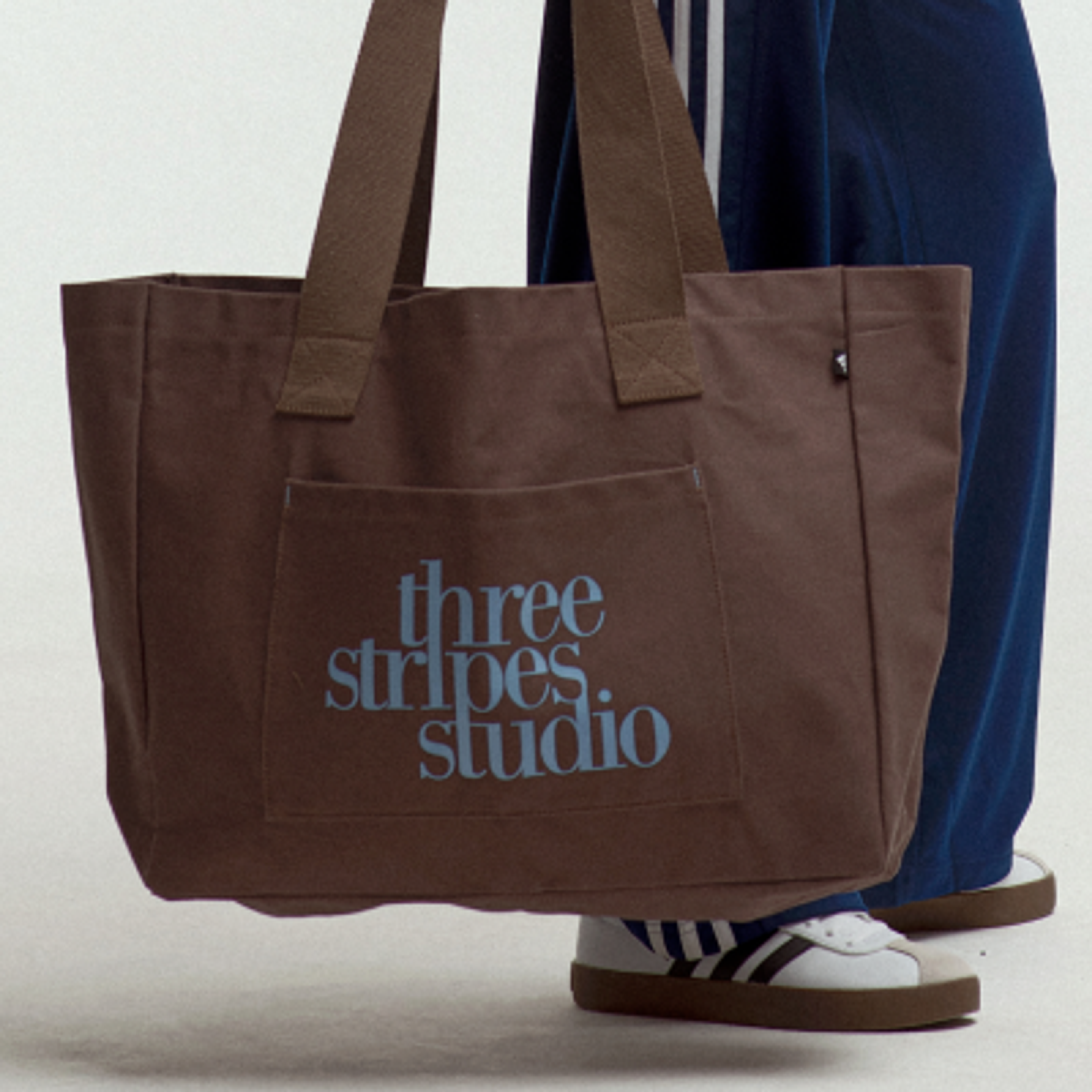 Brown canvas tote bag with "three stripes. studio." in light blue on a front pocket.
