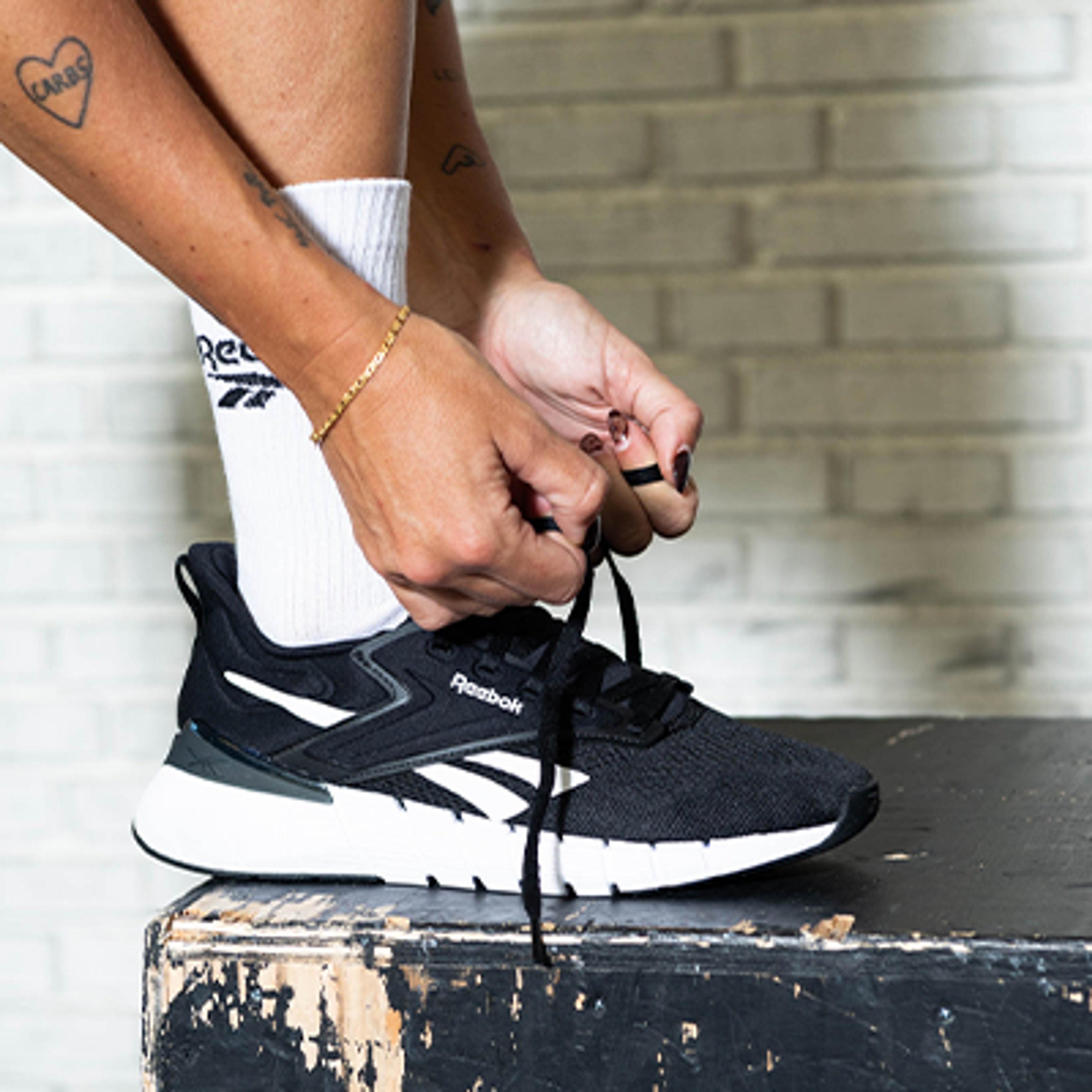 A person ties the laces of a black Reebok sneaker, wearing white Reebok socks.