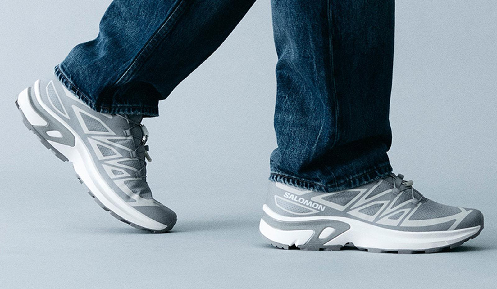 A person wearing blue jeans and light grey Salomon sneakers with white soles, mid-step.