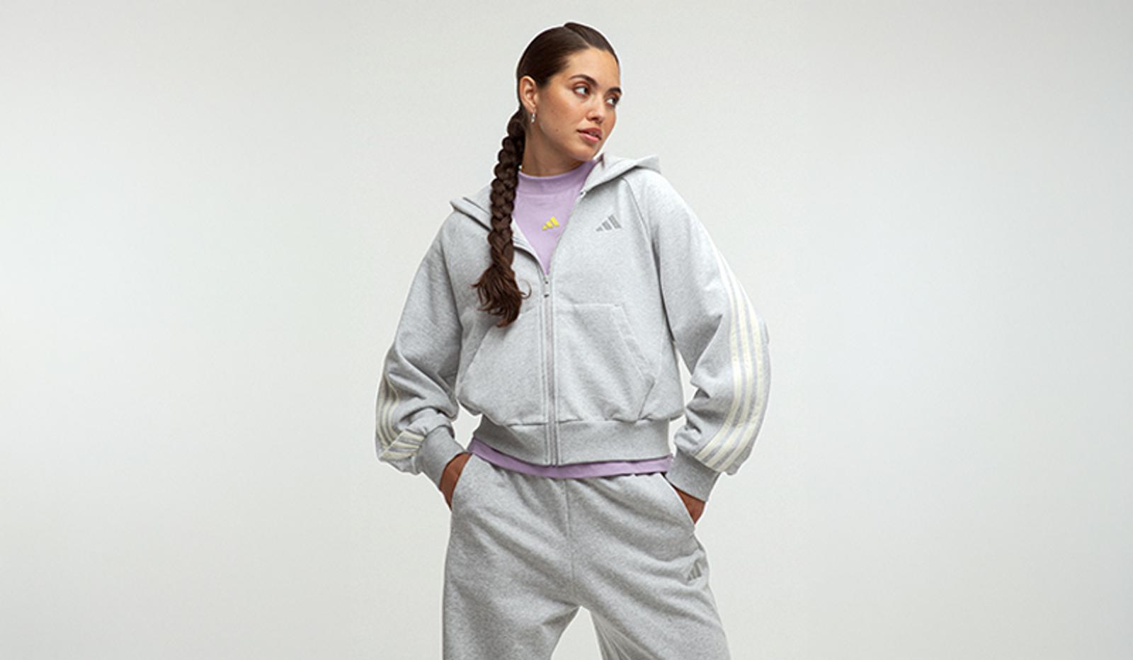 A woman wears a grey Adidas zip-up hoodie with three white stripes on the sleeves, matching sweatpants, and a purple top, looking to her right.