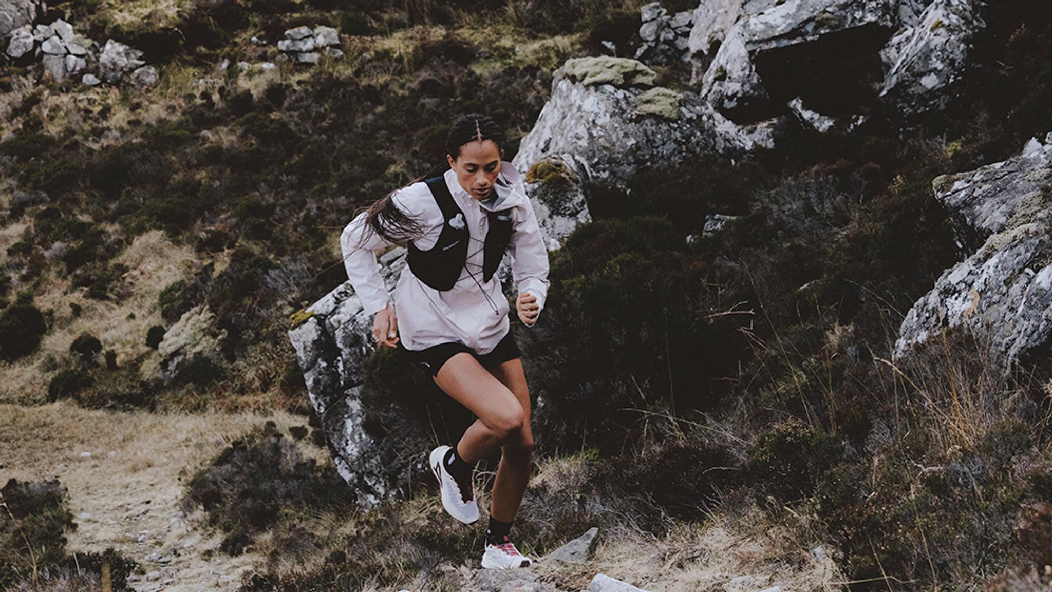 A woman trail running uphill on a rocky, brush-covered slope.