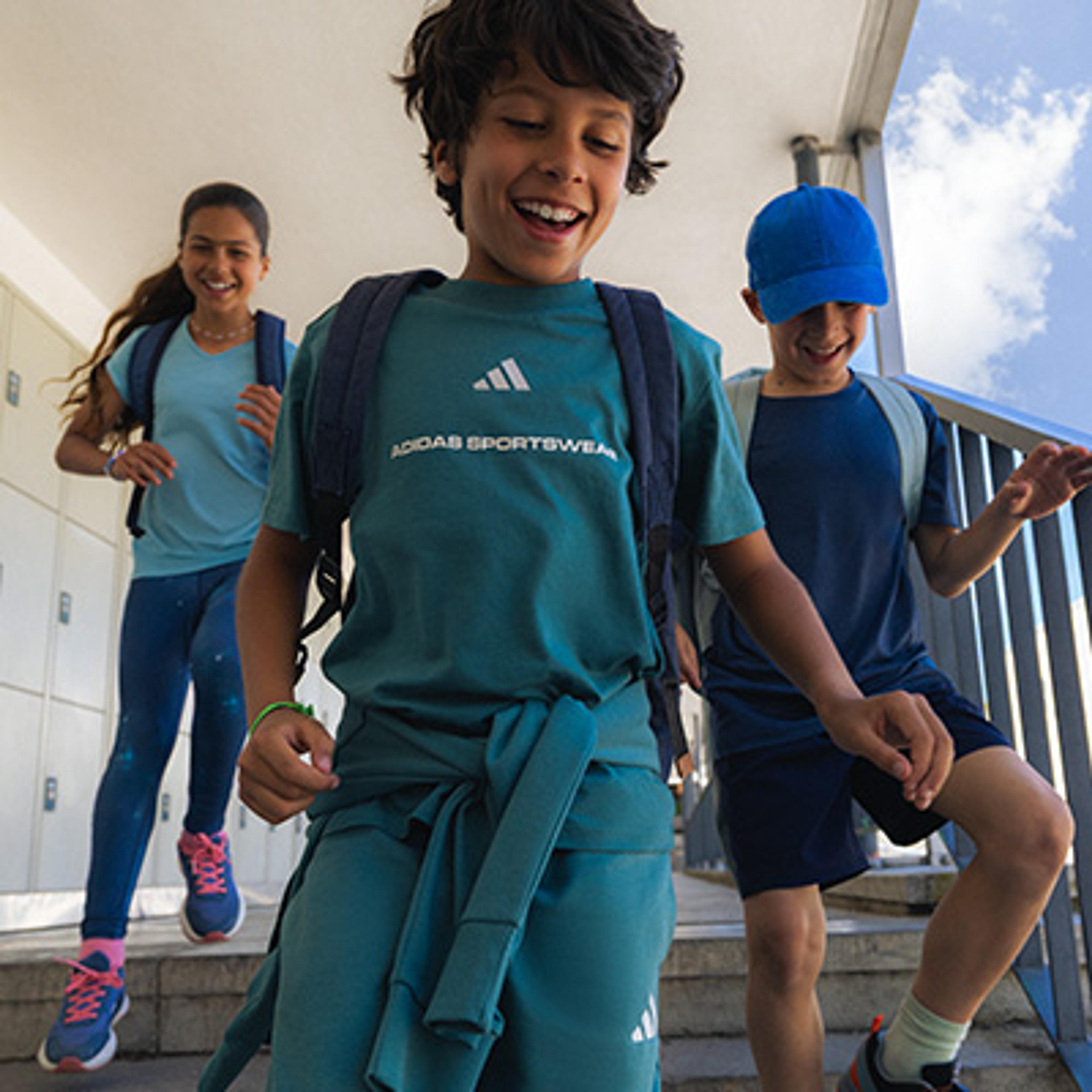 Three smiling children in athletic wear with backpacks walk down stairs.