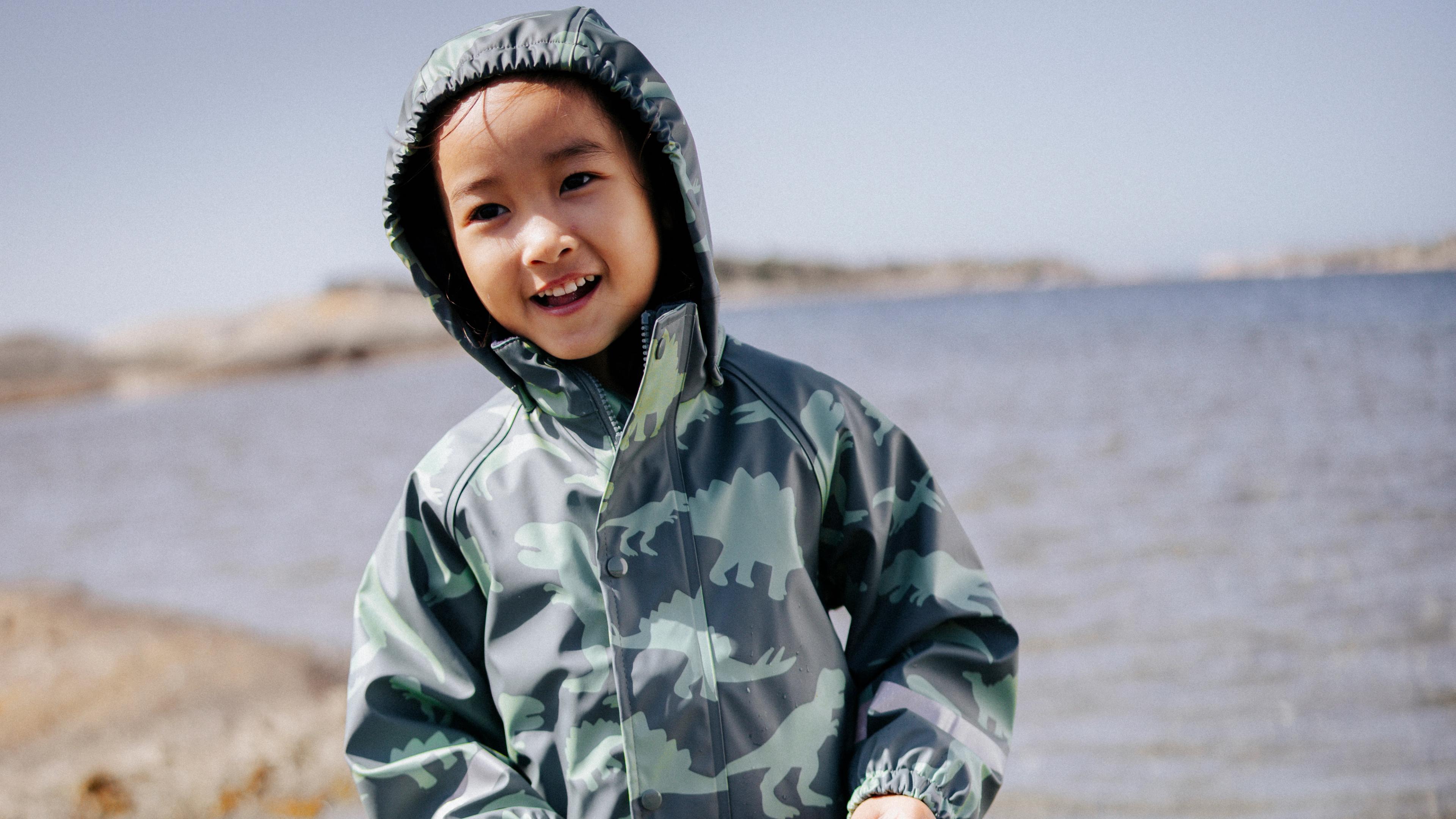 Smiling child in a hooded green dinosaur print rain jacket by the water.