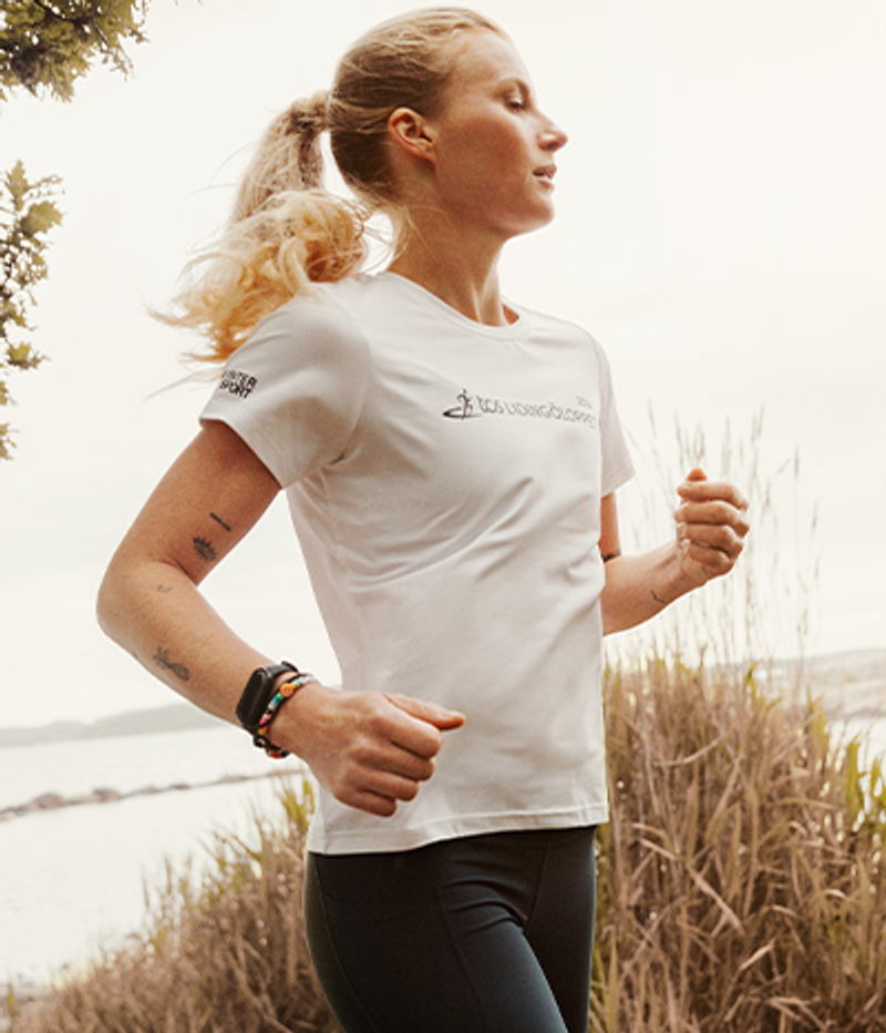 A blonde woman in a white running shirt with "TCS Lidingöloppet" branding runs outdoors by water.