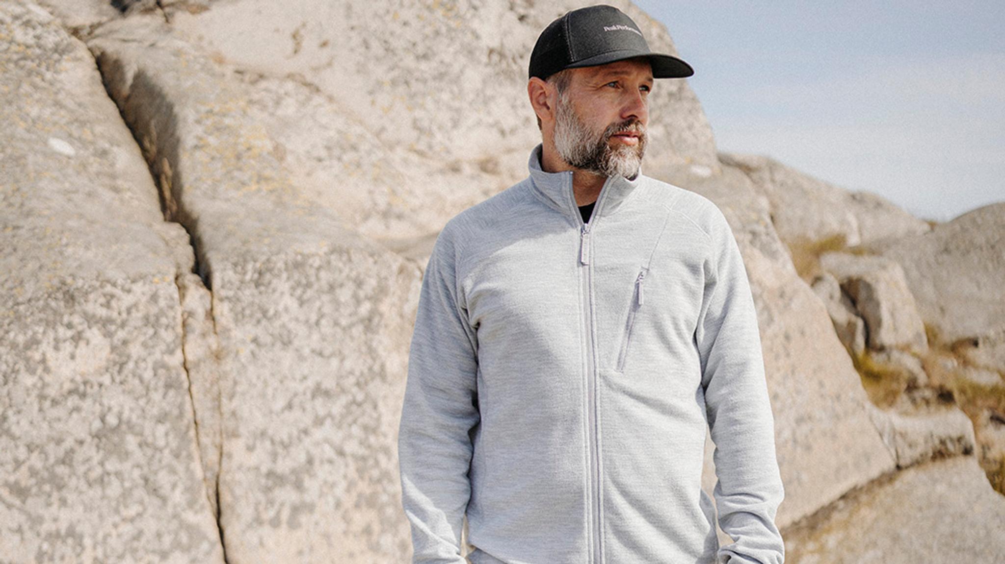 A bearded man in a gray zip-up jacket and black cap stands among light-colored rocks.