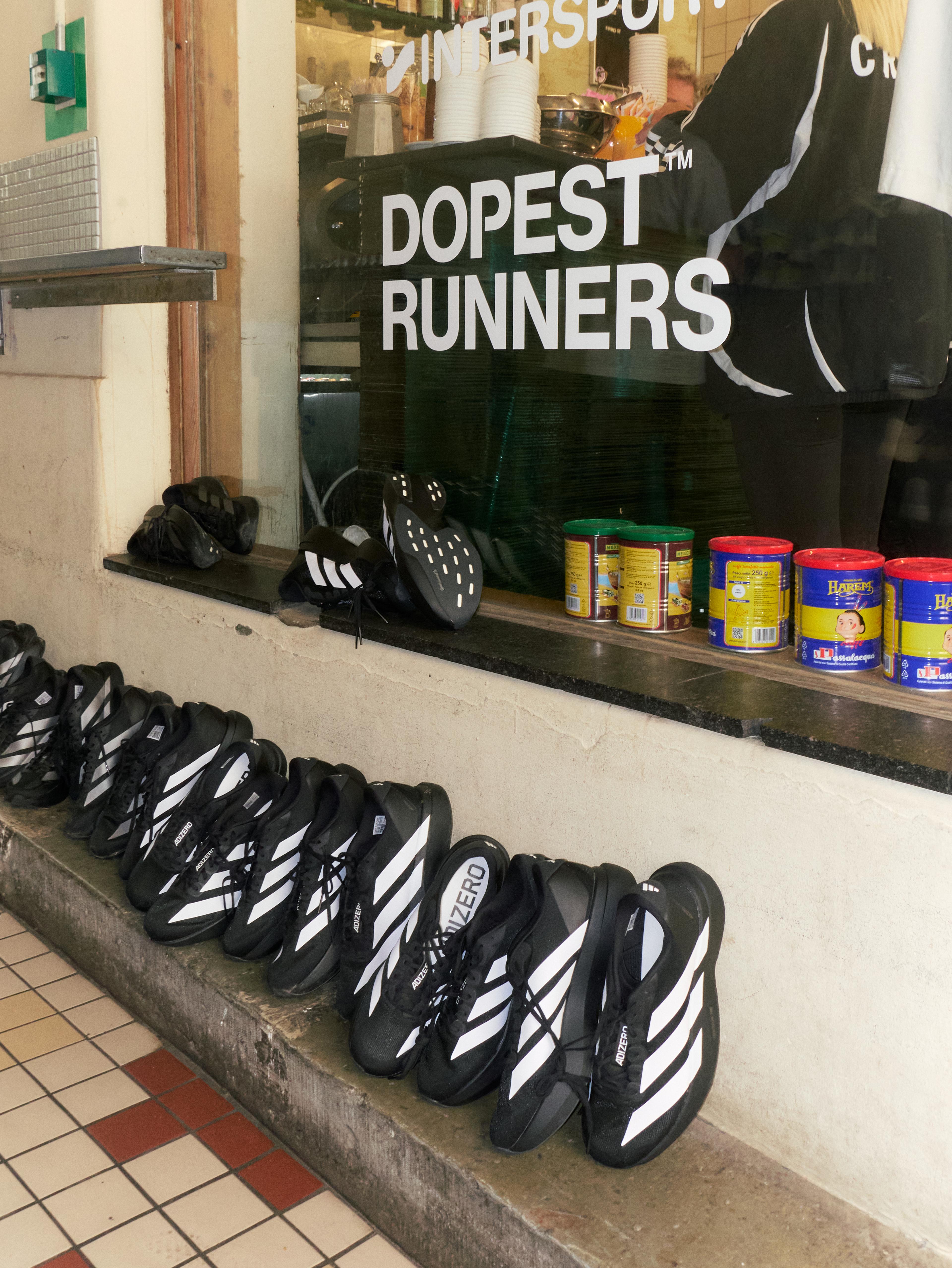 A row of black Adidas running shoes with white stripes displayed on a ledge outside a shop window that reads "DOPEST RUNNERS".