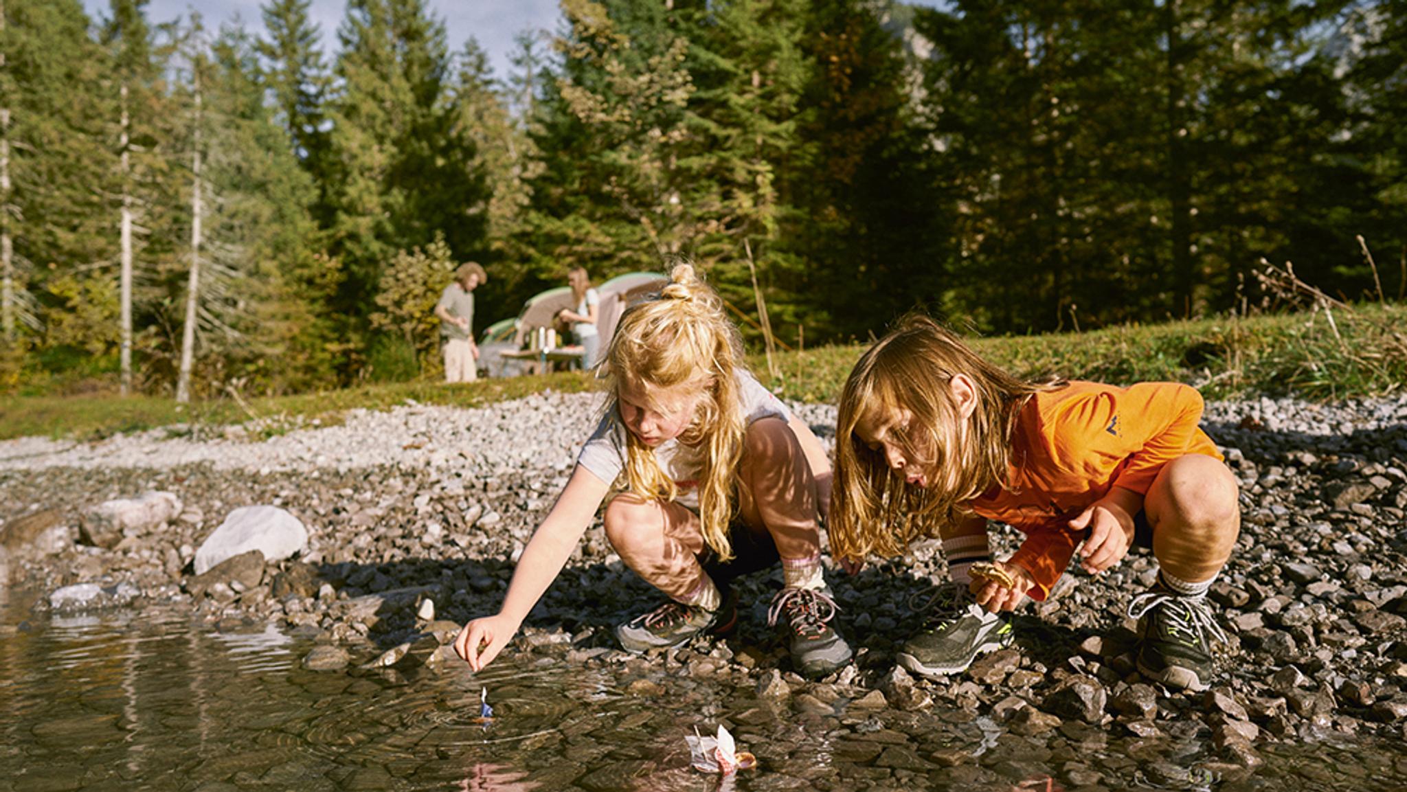 Two children play at the edge of a rocky lake, with a family campsite visible in the forest background.