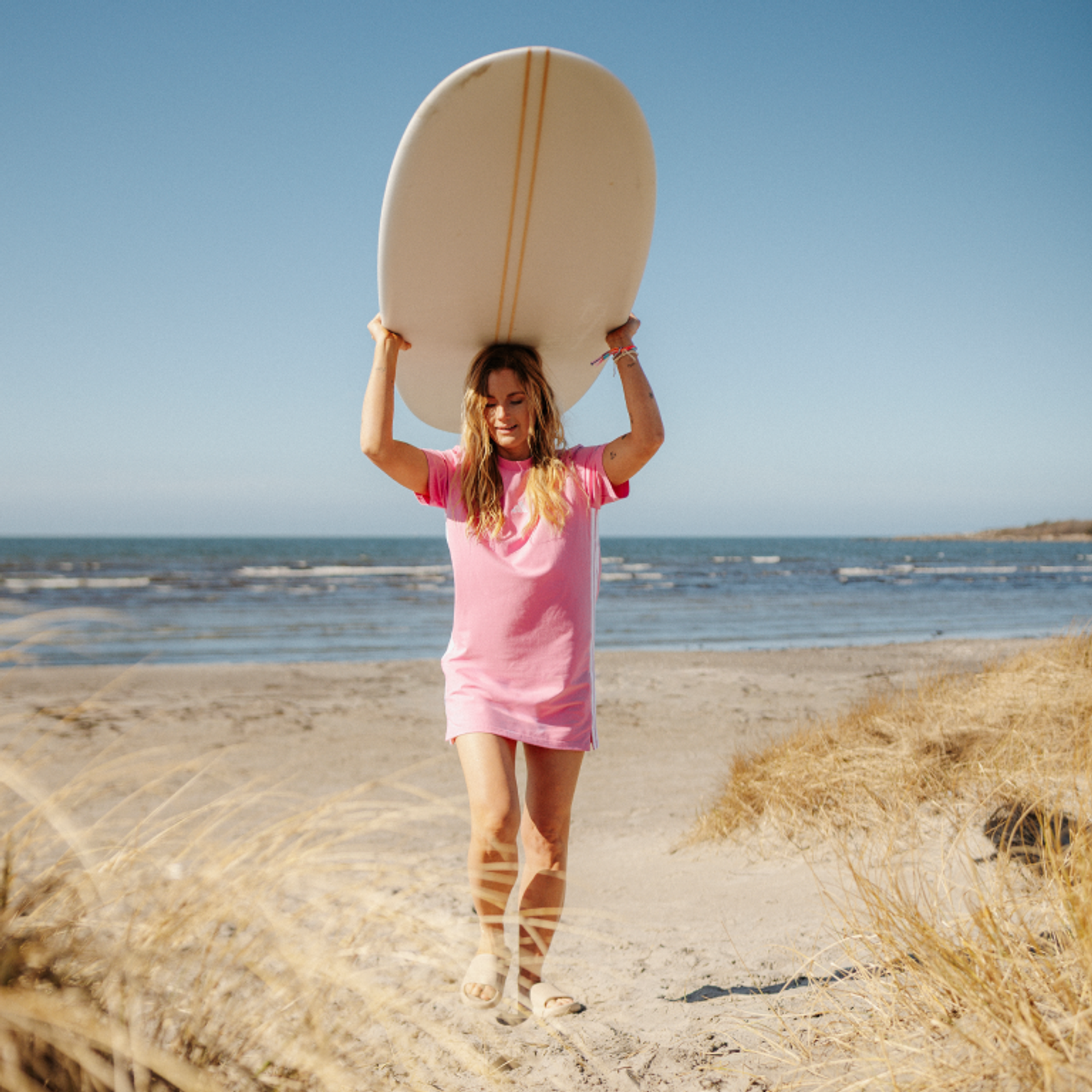 A woman in a pink dress walks on a sandy beach path, carrying a surfboard on her head.