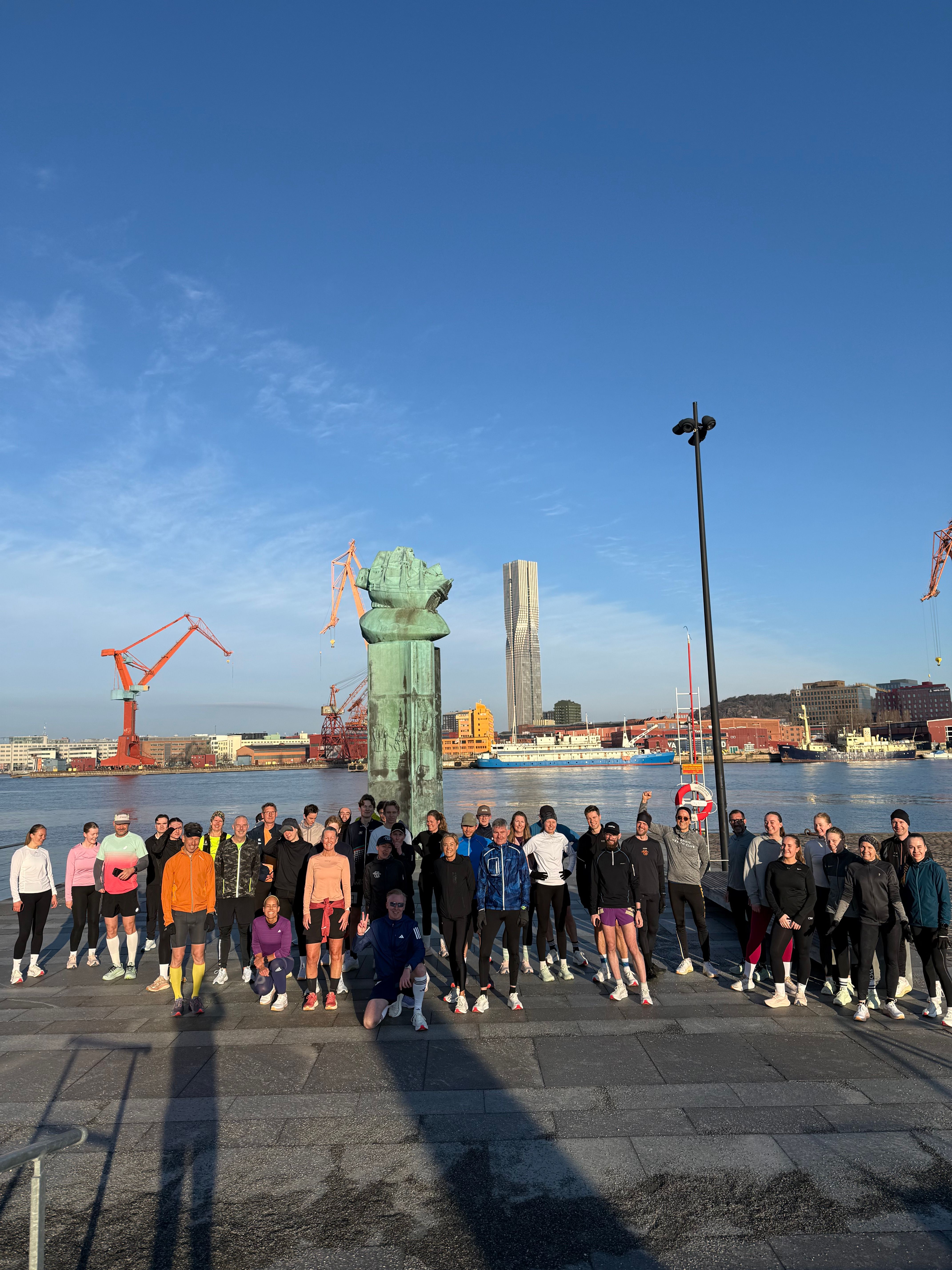 A large group of athletes in activewear pose by a waterfront with a large diver helmet statue, city buildings, and cranes in the background under a blue sky.