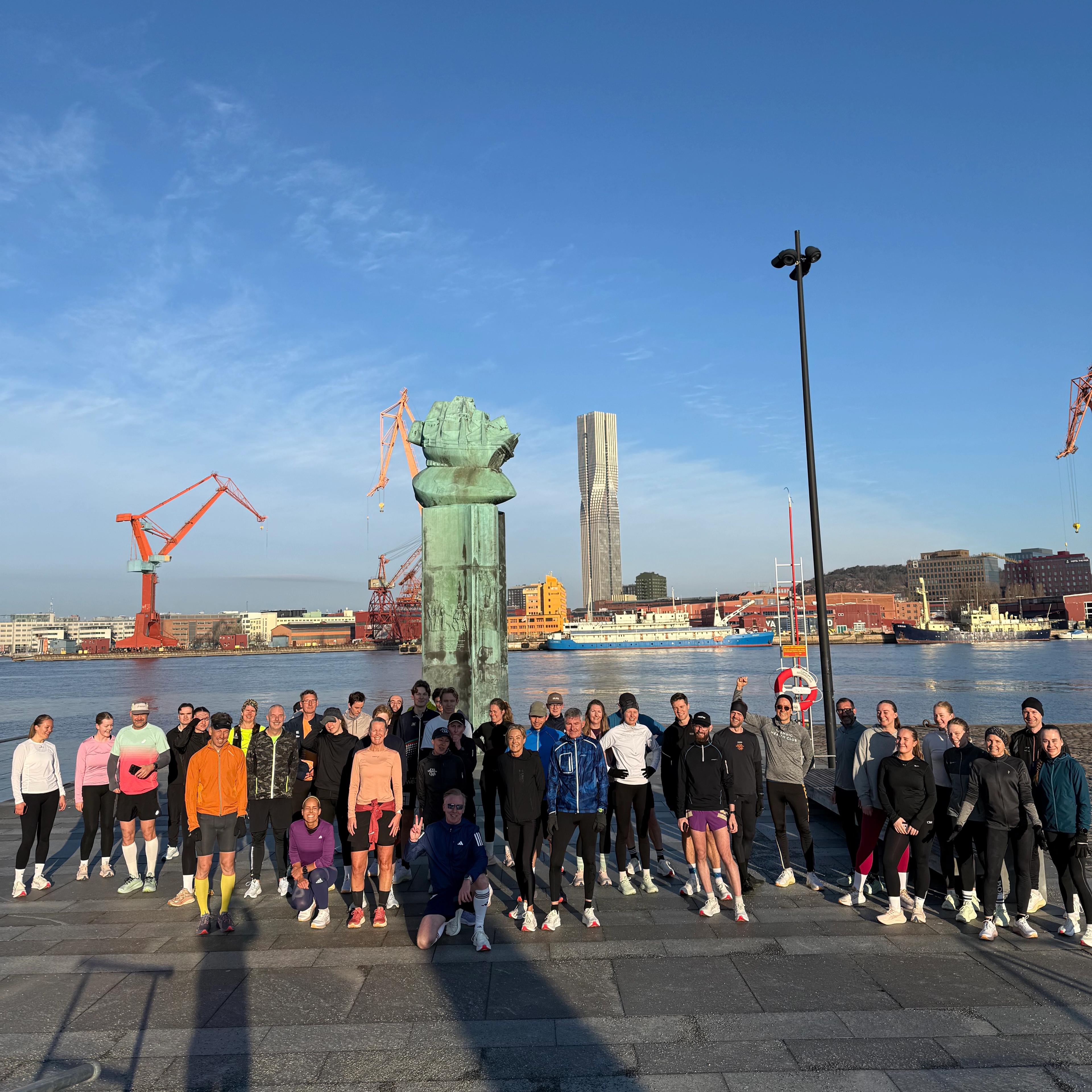 A large group of athletes in activewear pose by a waterfront with a large diver helmet statue, city buildings, and cranes in the background under a blue sky.